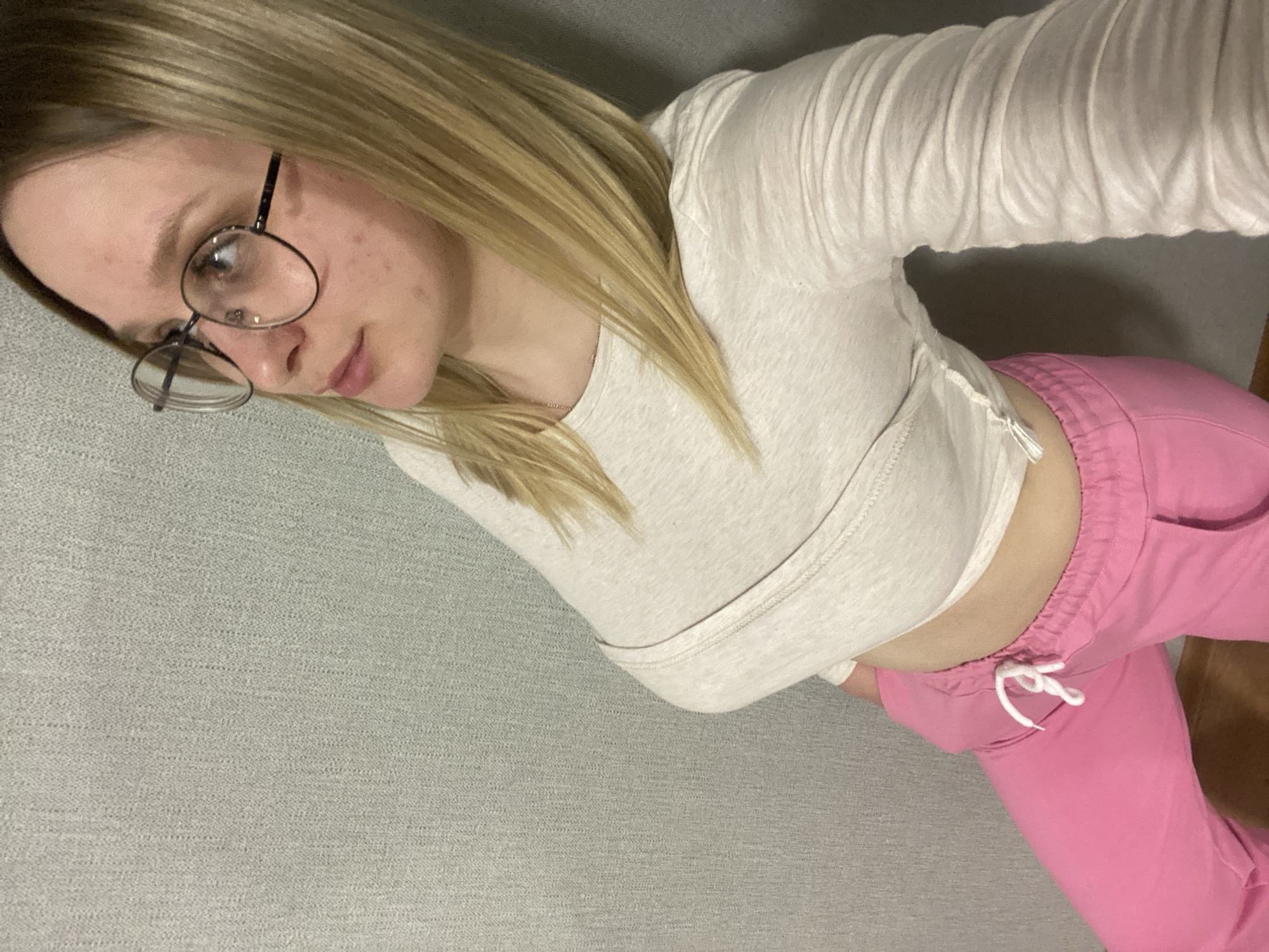 A fair-skinned woman with blonde hair and glasses is lying down. She is wearing a long-sleeved, light-colored shirt and pink pants. The background is a gray surface. The location cannot be determined from the image.