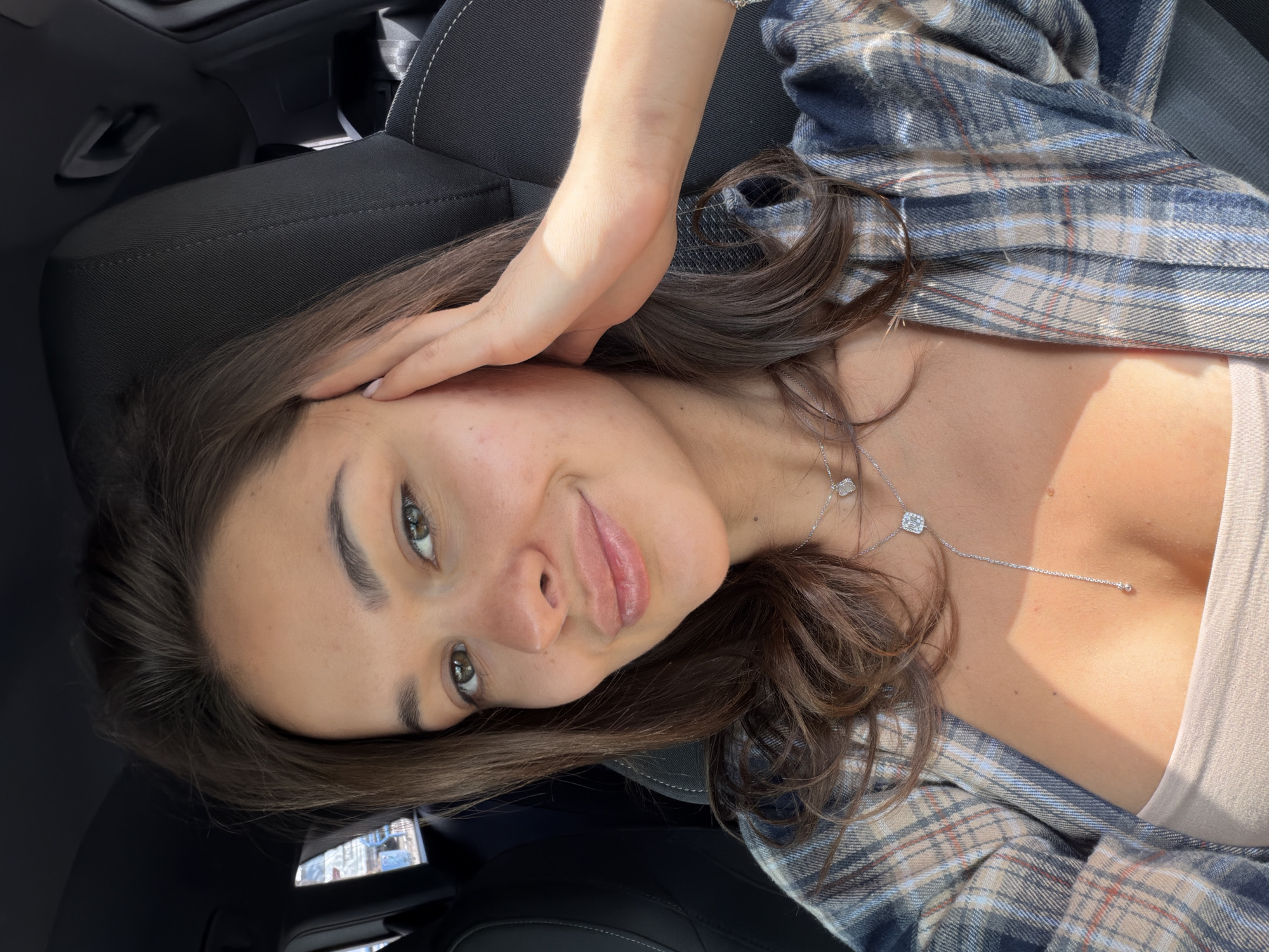 A woman is reclining in a car seat. She has brown hair, green eyes, and is wearing a plaid shirt over a neutral-colored top. A silver necklace is visible. She has her hand near her face and is smiling. The interior of the car is visible, including the black seat. There is no information to suggest that the location is in Krasnoyarsk, Russia.