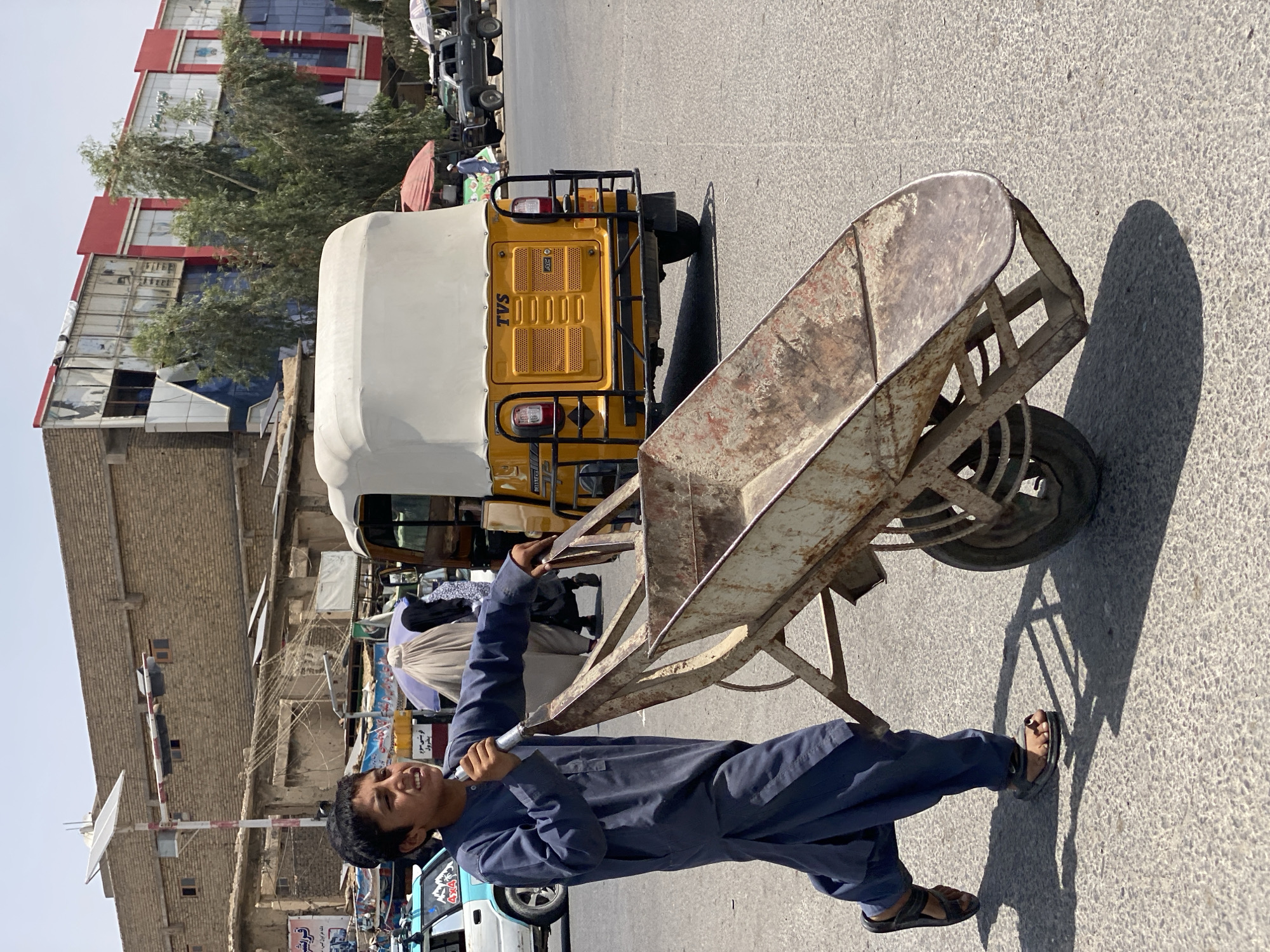 A young boy wearing a dark blue shalwar kameez and black sandals walks forward while pushing a rusty, metal wheelbarrow. He is on a paved road in an urban environment. Behind him, a bright yellow auto-rickshaw with a white canopy is visible, along with a white and blue car to the left. In the background, a multi-story building with a red facade stands amidst other structures and trees. The scene appears to be in a city, likely Kandahar, Afghanistan, given the architecture and vehicles. The lighting suggests it is daytime with bright sunlight casting shadows.