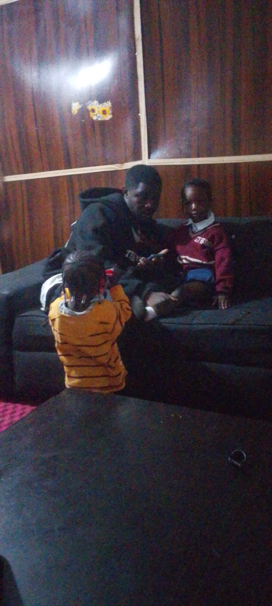 In an indoor setting with wood-paneled walls, a man in a black hooded sweatshirt sits on a dark couch, holding a mobile phone. Beside him, a young child in a maroon sweater and blue shorts looks towards the camera. In front of them, a child wearing a yellow and orange striped shirt is seated on the floor, facing away from the camera. The room is dimly lit, with a light source illuminating the upper portion of the wall where decorative floral stickers are visible. A dark, textured surface occupies the foreground.