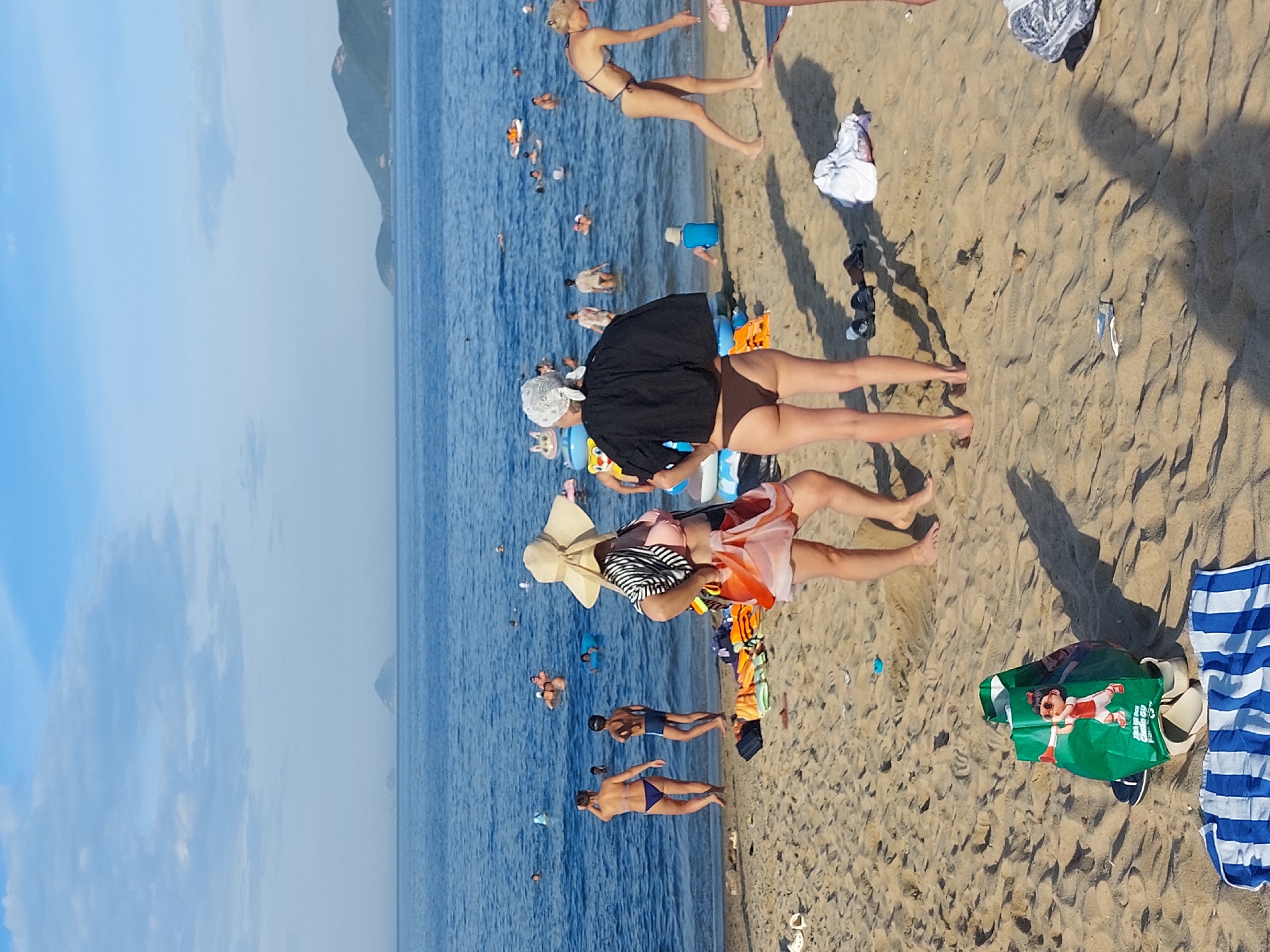 The image depicts a crowded beach scene in Nha Trang, Vietnam. The blue ocean stretches out to the horizon, with several people swimming and playing in the water. In the foreground, on the sandy beach, a few individuals are standing and walking. One person, wearing a wide-brimmed hat and a striped top with an orange skirt, is facing away from the camera. Next to them, another person, also facing away, is wearing a black top and brown bikini bottoms. Scattered on the sand are various beach items, including a green shopping bag with an image of a child, towels, and flip-flops. The overall atmosphere suggests a sunny day at a popular tourist destination. The presence of a shopping bag with Vietnamese text suggests the location is likely a public beach in Nha Trang.