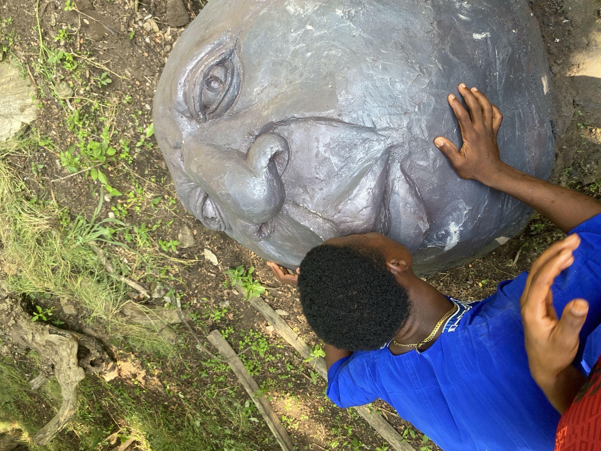 An overhead shot shows a person wearing a blue shirt and a gold chain leaning towards a large, gray, boulder-like sculpture of a face. The person is kissing the sculpture's lips, and their hands are resting on the sculpture. Another person's hand, wearing a red shirt, is visible in the lower left corner. The sculpture appears to be outdoors, on uneven ground with grass, dirt, and a fallen log in the background.