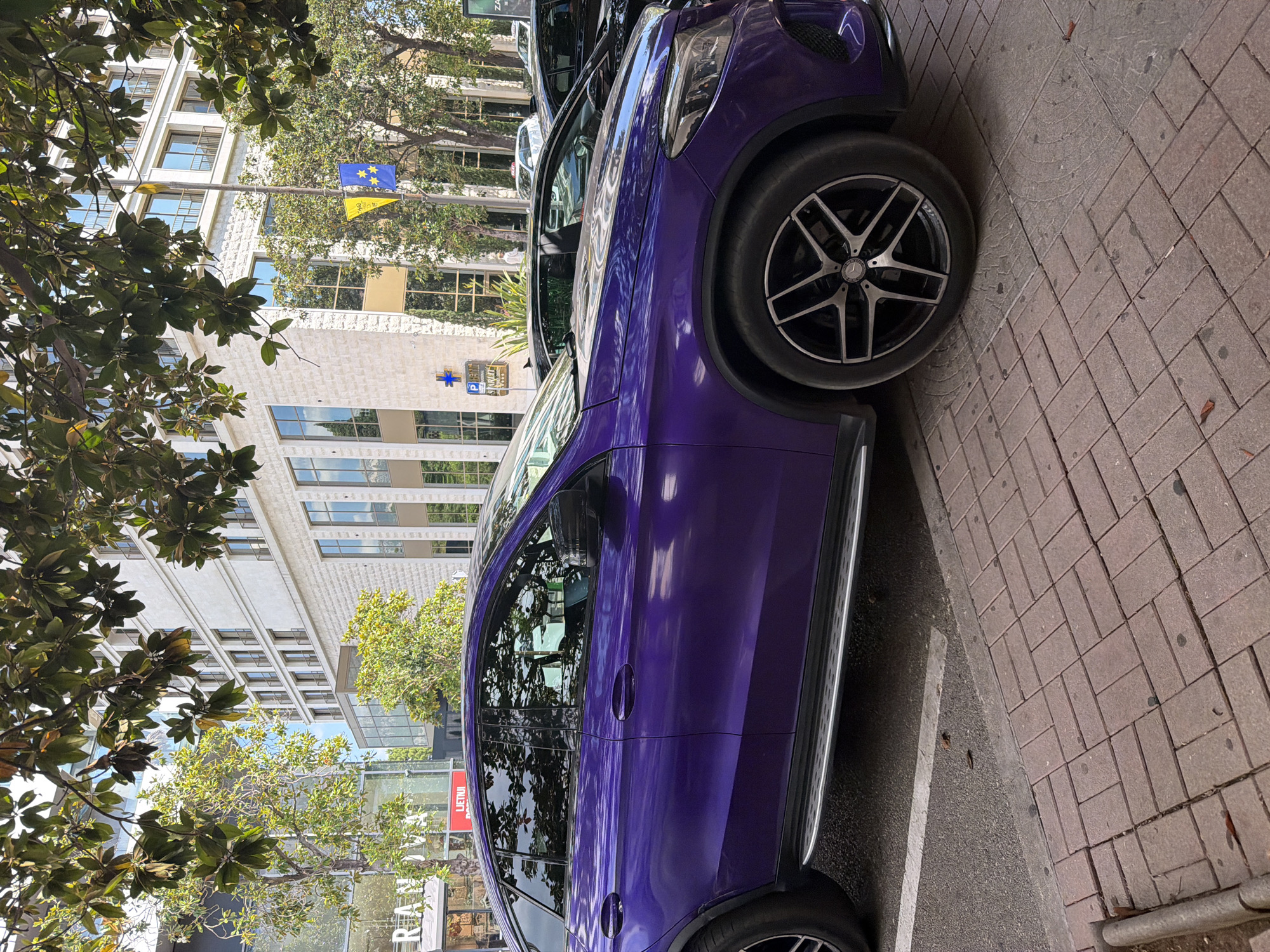 A purple Mercedes-Benz GLA is parked on a sidewalk paved with rectangular bricks. The car is the primary subject, with its sleek, metallic finish reflecting the surrounding environment. Part of a second car, also dark-colored, is visible behind the Mercedes.
The setting is an urban environment, likely a street in Budva, Montenegro, given the context. Behind the cars, a modern, multi-story building with a white facade and numerous windows stands. Trees with lush green foliage are visible in the foreground and between the cars, casting shadows on the sidewalk and the car's surface. One tree's branches are prominent in the upper left corner of the image.
A traffic light pole is present, with two small flags hanging from it: one blue and yellow, and another yellow with some symbols. A blue sign with a white cross and a white "P" inside a blue circle, likely indicating parking, is affixed to the building.
The time of day appears to be daytime, with sunlight creating highlights and shadows, suggesting fair weather. The image is taken from a low angle, focusing on the car's side profile and its wheel. The overall mood is calm and ordinary, capturing a moment of a parked car in a city setting. There is text visible on a sign attached to a building in the background, which reads "LIETUVAI".