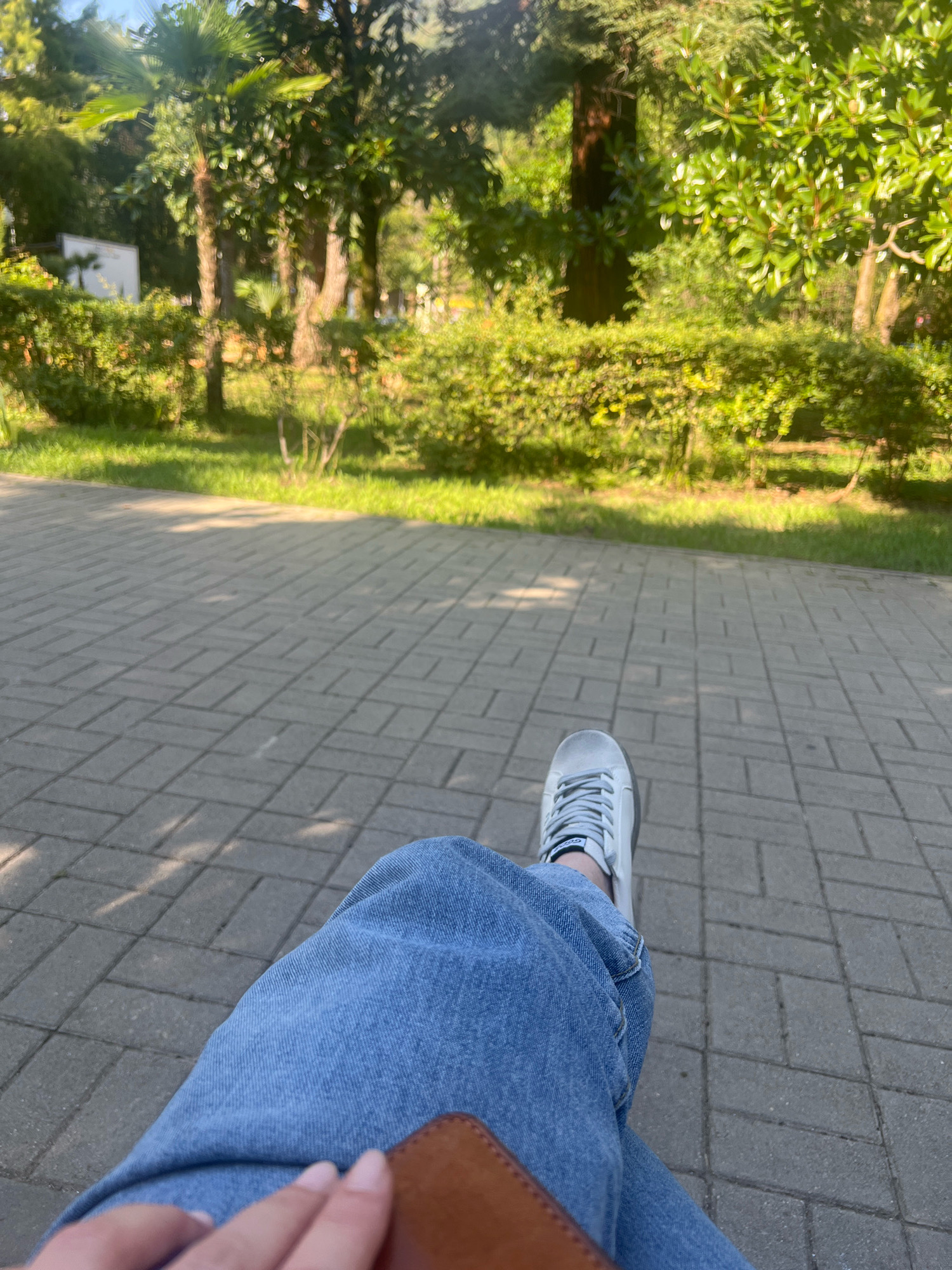 A first-person perspective shot shows a person's legs and feet resting on a paved area. The person is wearing blue jeans and white sneakers. In the background, there is a lush green park with trees and bushes. The sun is casting shadows on the ground, indicating it's daytime. The location is identified as a park in New Athos, Abkhazia.