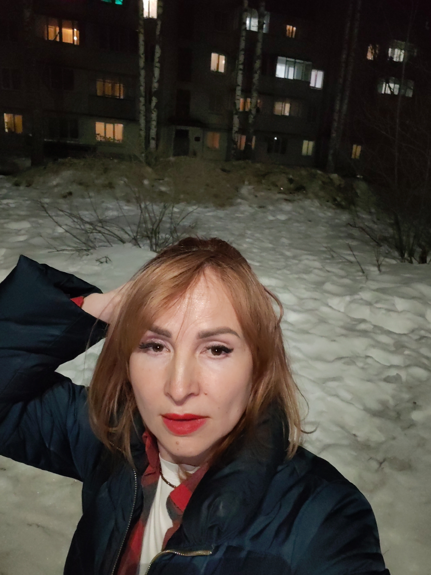 A woman with reddish-brown hair stands outdoors in front of a snow-covered landscape. She wears a dark blue puffer jacket over a white t-shirt and red plaid shirt. Her makeup includes red lipstick and dark eye makeup. Her right hand is raised and resting on the back of her head. Behind her is a multi-story building with illuminated windows. Bare trees stand between the building and the woman. The setting is most likely the city of Chaikovsky, Russia, during winter.
