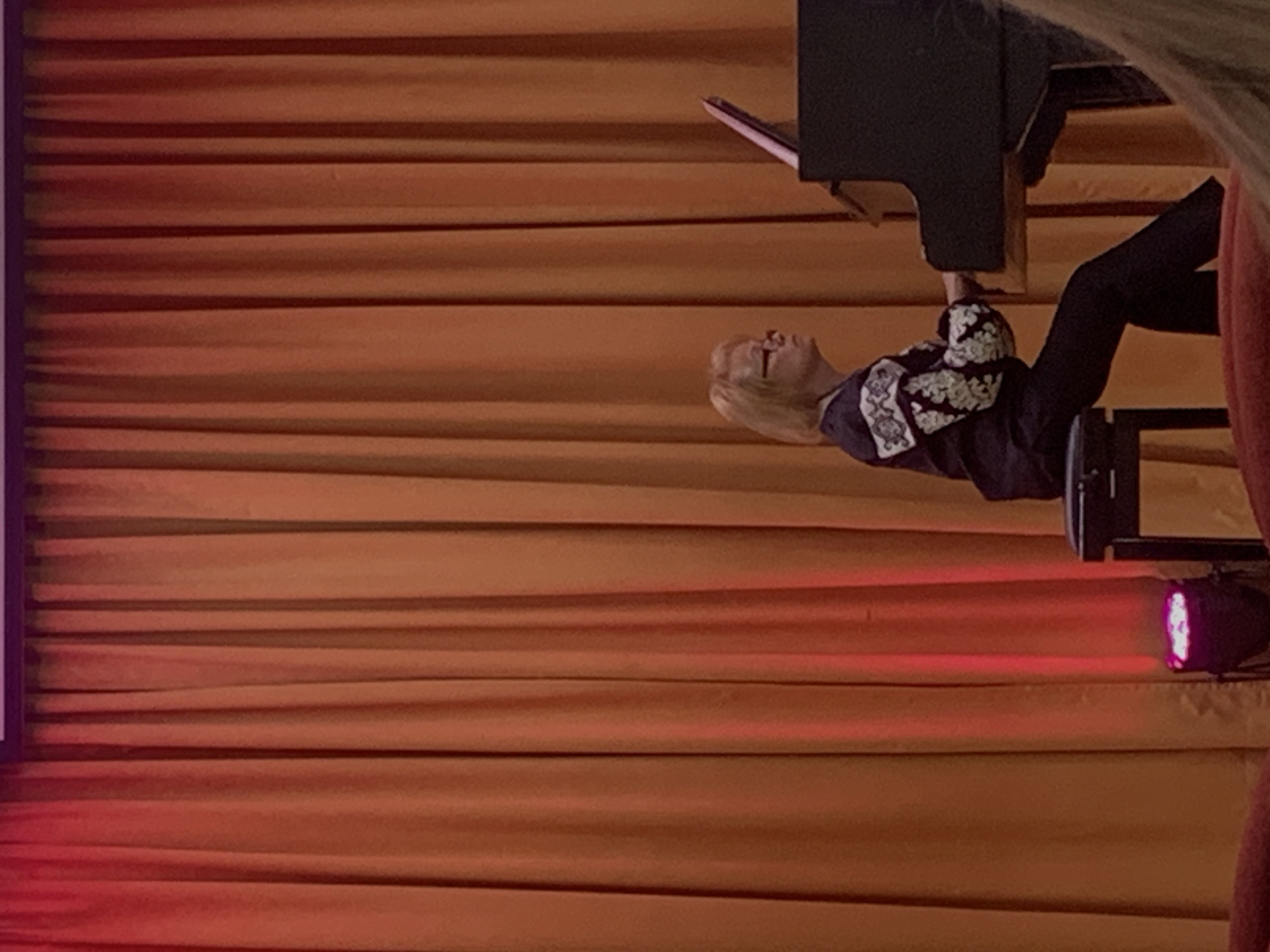 A woman with blonde hair and glasses plays a piano on a stage in front of an orange curtain. She is wearing a dark blue shirt with embroidered white details on the sleeves and shoulders, and dark pants. The piano is black, and a music stand is attached to it. A stool is placed under the piano bench. A pink and purple stage light is visible on the floor at the bottom left of the image. The overall setting suggests a performance or recital venue. The specific location in Kyiv, Ukraine cannot be determined from the image alone.