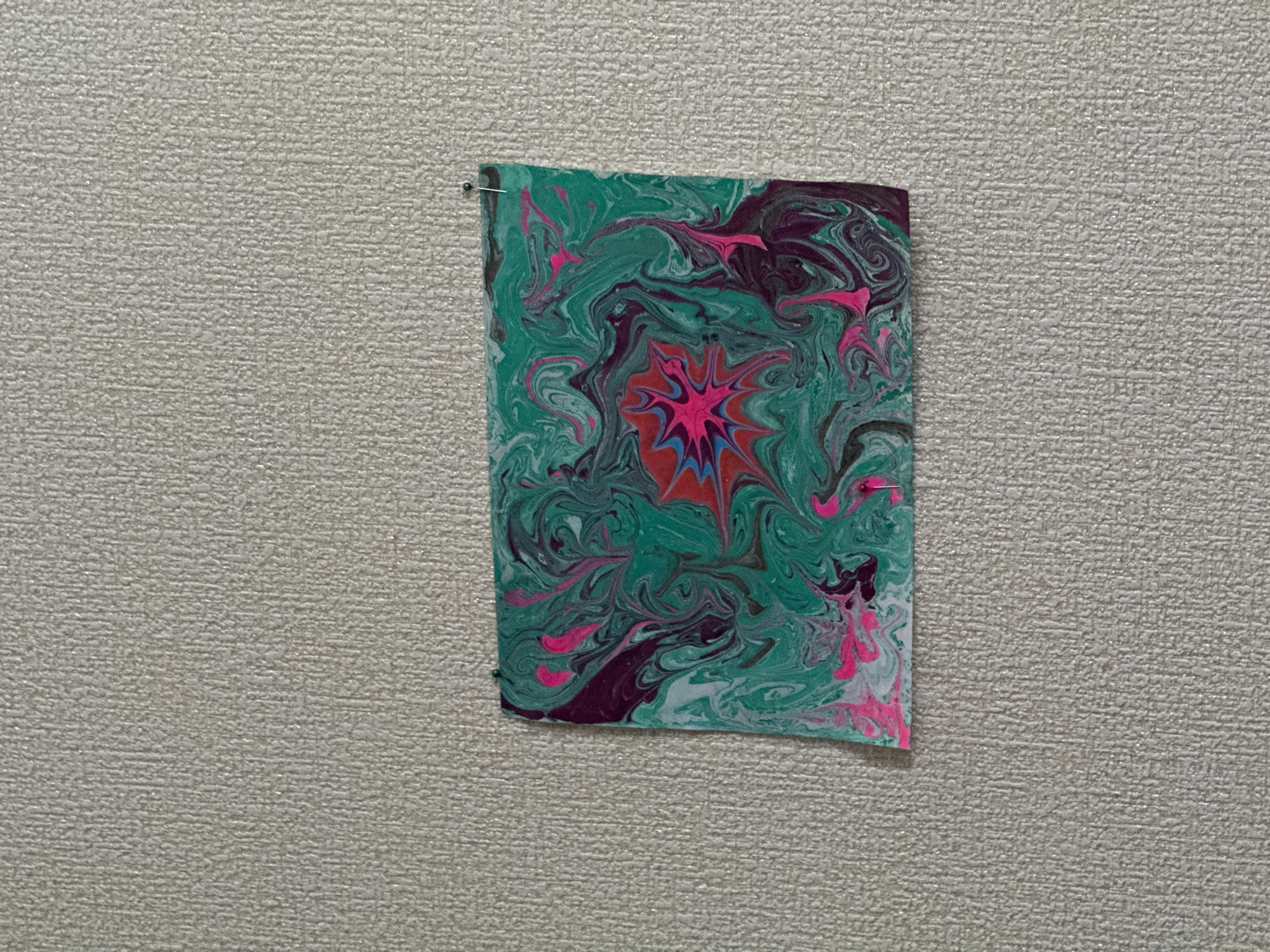 A rectangular piece of paper is pinned to a textured wall. The paper features a marbled design of green, pink, red, purple, and white colors. The design appears to have a central, starburst-like pattern of red, pink, and blue hues. The wall has a light-colored, speckled texture. The location is Unknown, Unknown.