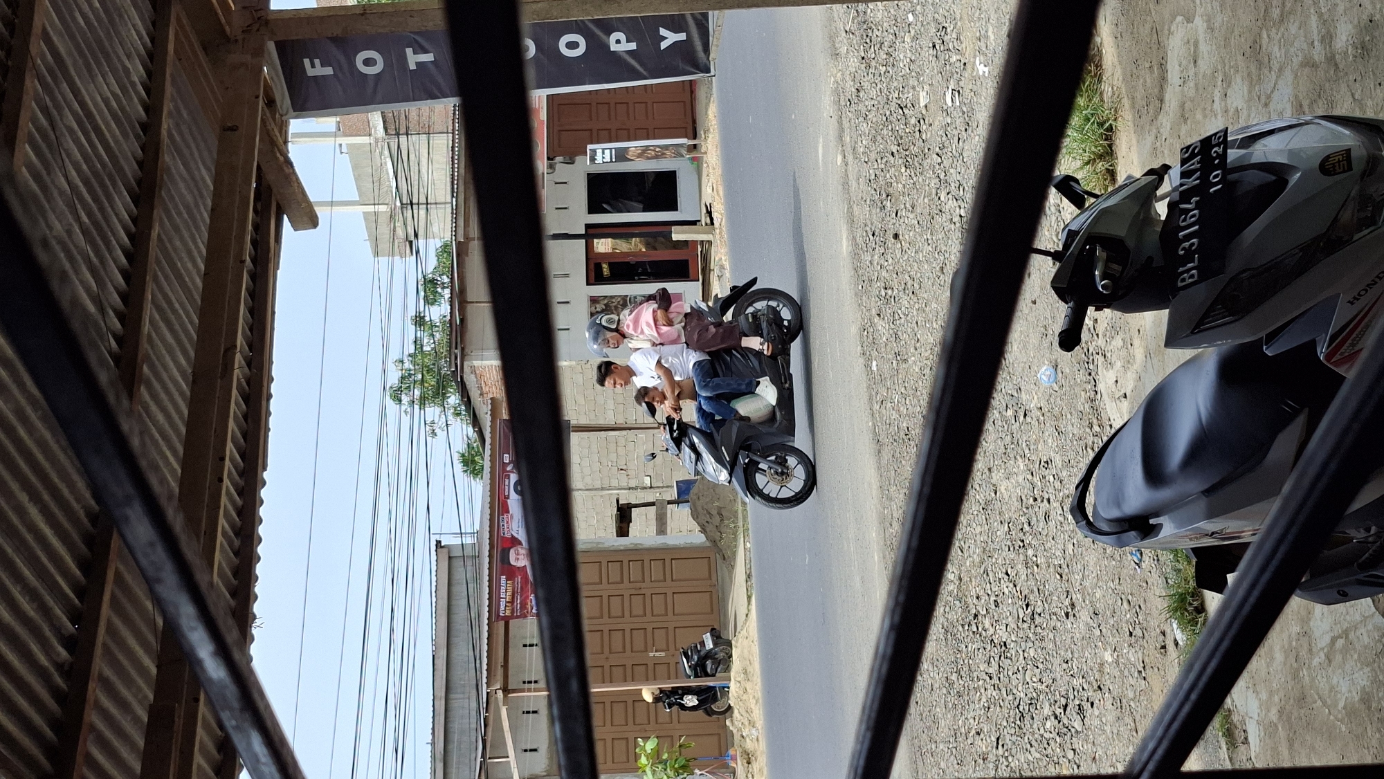 In Keudebayu, Indonesia, a family of three rides a black motorcycle along a paved road. The father, wearing a white t-shirt and blue jeans, drives the motorcycle. His wife, in a pink hijab and brown pants, sits behind him, holding their child. The child, wearing a blue shirt, is seated between them. A dark-colored scooter is parked on the side of the road to the left. Buildings with corrugated metal roofs line the street, and a banner with the word "FOTCOPY" is visible on the right. The ground is covered with gravel.