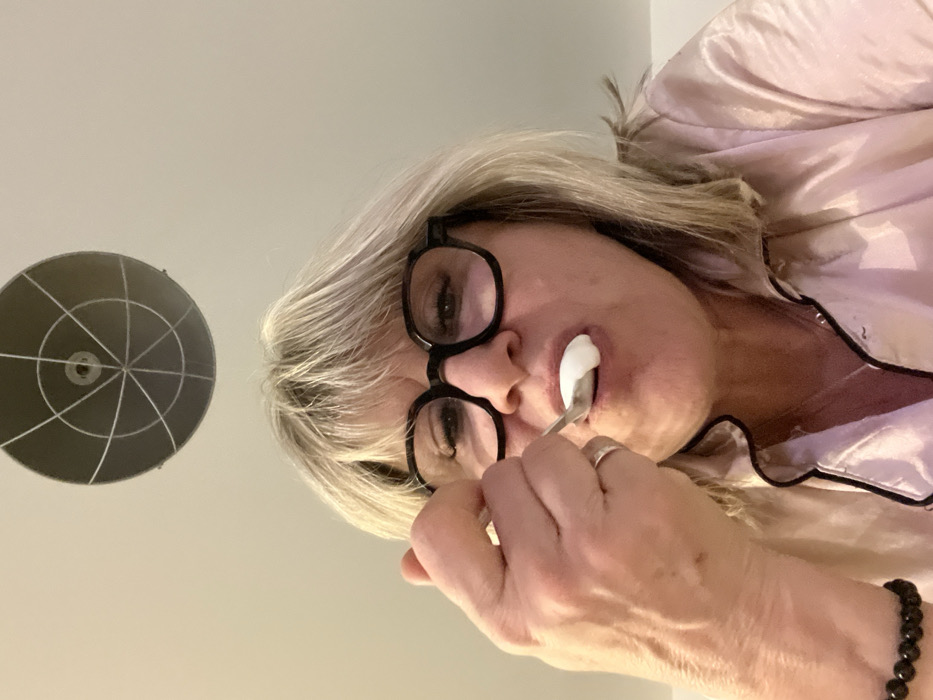 The image presents a close-up, angled shot of an adult individual, likely a woman, with light-colored hair, possibly blonde or gray. The person is wearing black-framed glasses and appears to be in the process of consuming a white, frothy substance from a small metal spoon. There is some of the white substance visible on their lower lip and chin area.
The individual is dressed in what looks like a light pink, satin or silk-like pajama top with dark (possibly black) piping around the collar and front opening. A silver-toned ring is visible on the ring finger of their left hand, which is holding the spoon. A dark, beaded bracelet is partially visible on their right wrist.
The background is a plain, light-colored surface, presumably a ceiling or wall. In the upper left portion of the image, there is a distinct circular object mounted on this surface. It appears to be a light fixture or a ventilation grate, featuring a central point with several radial lines extending outwards, creating a spoke-like or target pattern.
The overall perspective is from a low angle, looking upwards at the person, or the image itself is rotated, making the person's head appear significantly tilted to the right. The lighting is soft and even. No text is visible in the image.