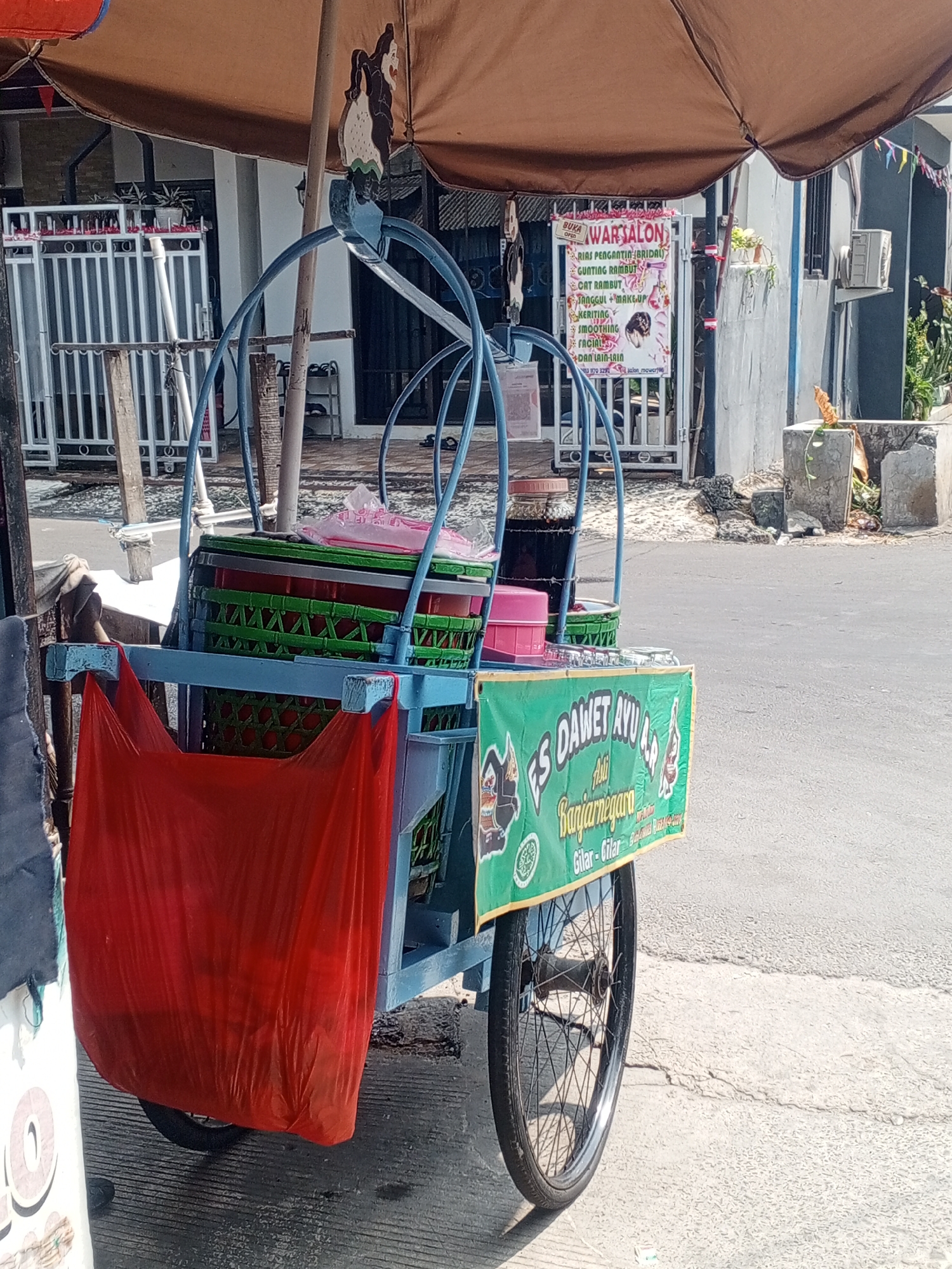 An outdoor, eye-level, medium shot shows a street vendor's cart parked on a cracked concrete surface under a brown canopy in Depok, Indonesia. The cart, painted blue, has a large red plastic bag hanging from its front, obscuring its lower portion. Above the bag, a green banner is affixed to the cart, displaying the words "ES DAWEL AYU" in large, white, bubble letters, followed by "Asli Banjarnegara" and smaller text indicating locations such as "Gilar-Gilar" and "Bumiayu-Banjar". The cart contains several baskets, some filled with green and red layered items, and glass containers with dark liquid. A wooden decorative figure is mounted on the top left of the cart's frame. In the background, a white fence with metal bars stands in front of a building with a gray wall, and a silver air conditioning unit is visible. The sun casts shadows, indicating daylight.