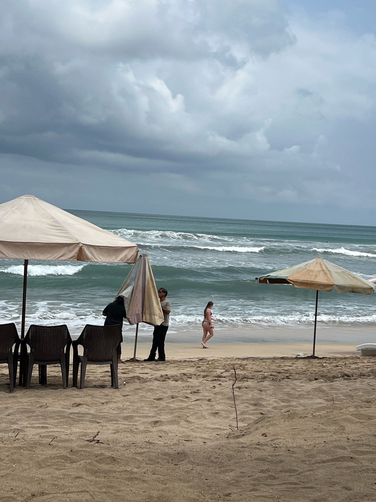 This image depicts a cloudy day at Kuta Beach in Bali, Indonesia. In the foreground, there are several dark, woven plastic chairs arranged on the sandy beach. Behind the chairs, two beach umbrellas stand. One large, light-colored umbrella is to the left, and a smaller, weathered umbrella with striped fabric is in the center. Two individuals are near the smaller umbrella, one appearing to be adjusting it. To the right, a person in a bikini walks along the wet sand near the water's edge. The ocean has moderate waves crashing onto the shore. The sky is filled with heavy, gray clouds, indicating potential rain. The overall scene suggests a somewhat overcast but still active beach environment.