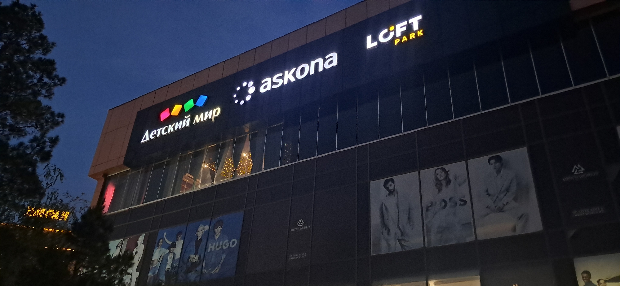 An exterior shot of a modern, multi-story building at dusk. The building's facade is dark, with large glass windows reflecting the twilight sky. Illuminated signage is prominently displayed across the top of the building. From left to right, the signs read "Детский мир" (Detkiy Mir), a Russian children's clothing store, followed by "askona," and finally "LCFT PARK." Below the upper level signage, the second floor also features illuminated advertisements. One prominent advertisement displays "HUGO." Further down, on the third floor, several large monochrome billboards feature fashion models. One billboard shows a male model wearing a suit, and another displays a female model wearing an outfit with the "BOSS" logo. Another billboard shows a male model in a suit. The scene is illuminated by the building's internal lights and external signage, creating a nighttime ambiance. A tree is visible on the left side of the frame, partially obscuring the building.