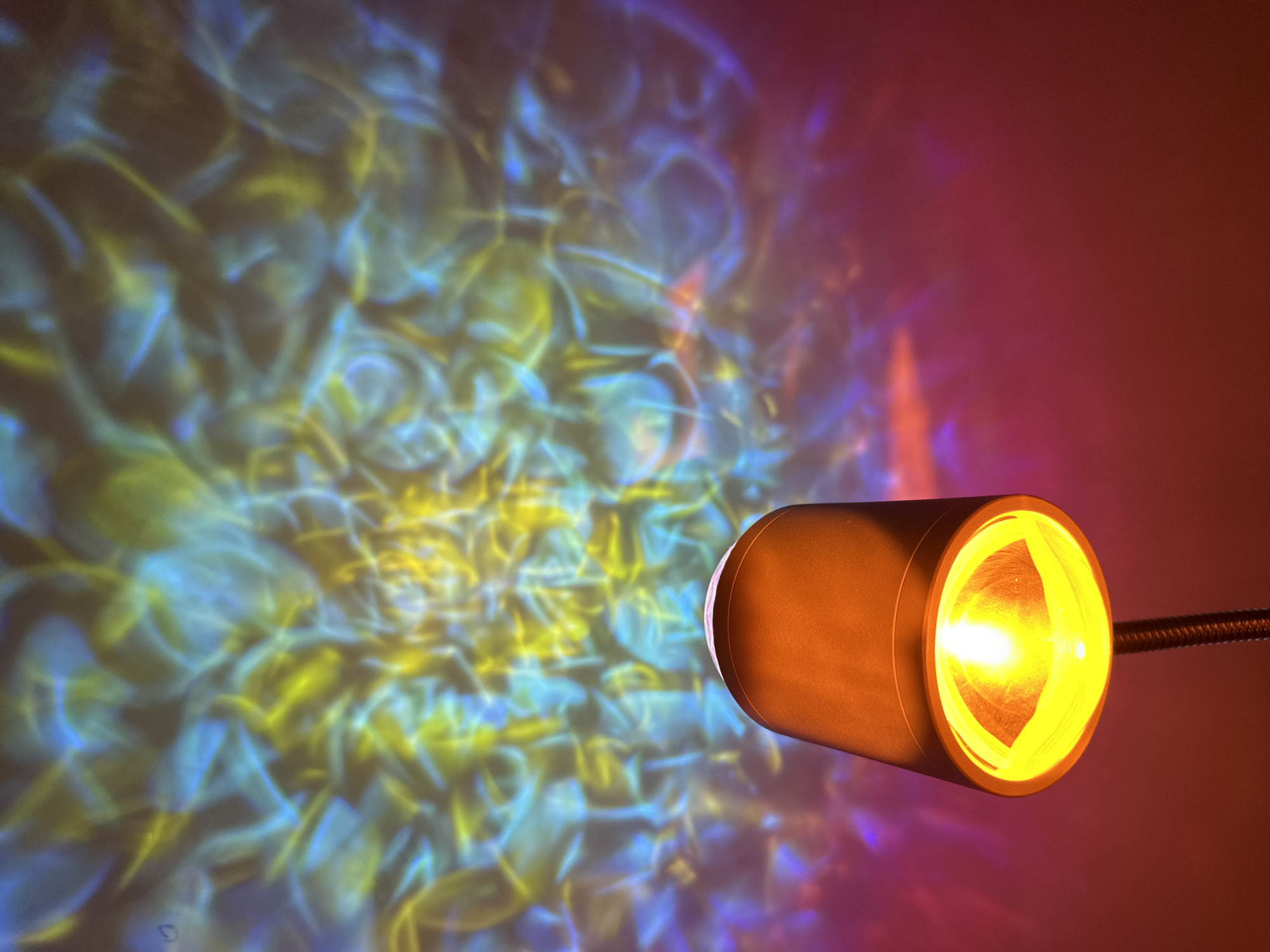 The image shows a cylindrical orange lamp illuminating a wall. The wall displays a pattern of diffused blue and yellow light, creating a water-like effect. The lamp is positioned on the right side of the image, emitting a bright yellow glow. The location is unknown.
