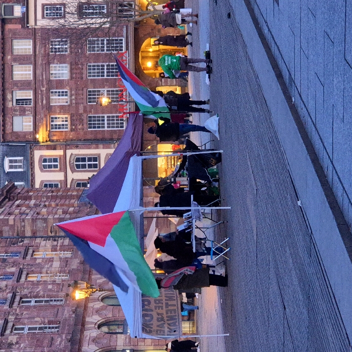 In a European city, a group gathers, displaying Palestinian flags. They are situated on a brick-paved area near buildings with distinct architecture. A sign is visible, though the text is partially obscured. Some individuals are under tents, while others stand. One person wears a bright green hooded jacket. The atmosphere appears to be a political gathering or demonstration.