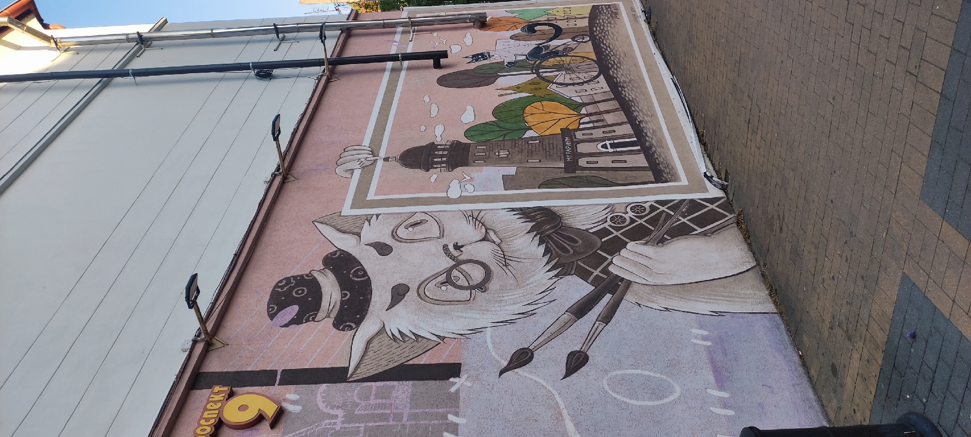 The media file displays a vibrant mural painted on the side of a building, next to a paved pedestrian area, likely in Zelenogradsk, Russia. The mural's dominant feature is a whimsical, anthropomorphic cat-like character depicted as an artist. This creature has white fur, wears round glasses and a patterned hat, and appears to have a monocle on its right eye. It holds three paintbrushes in its paws, suggesting an act of creation.
Above the cat, within the mural, a framed scene unfolds. It shows a coastal town setting with several buildings, including a prominent lighthouse or tower, and leafy trees in various shades of green and yellow. A person is seen riding a bicycle in this background scene. The architectural elements and the text "МОР ПАРАД" (Sea Parade) visible on one of the buildings within the mural suggest a maritime theme, fitting for a coastal town like Zelenogradsk.
The building itself has a light-colored, possibly off-white, paneled facade on the left, with dark pipes running horizontally across its upper section. Several small black floodlights are affixed to the building, pointing down onto the mural, indicating it might be illuminated at night. The ground adjacent to the mural is a paved walkway made of interlocking dark gray and brown bricks.
On the lower left of the mural, partially visible text in yellow Cyrillic reads "ПРОСПЕКТ" (PROSPEKT) and a large number "9". The scene is captured during the daytime under a clear blue sky, with bright, even lighting illuminating the mural and surrounding area.