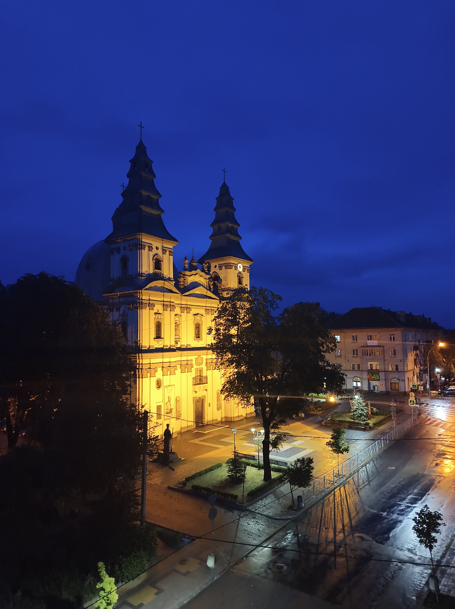 An exterior, eye-level, long shot captures a dimly lit Ternopil, Ukraine city square at dusk. The sky is a deep, dark blue with scattered clouds. On the left, a large, ornate, brightly lit church with two prominent spires dominates the scene. The church is illuminated by warm yellow lights, highlighting its architectural details. In front of the church, a dark silhouette of a statue stands on a pedestal.
To the right of the church, a wet asphalt road reflects the lights from the church and street lamps. Several trees are scattered throughout the square, some with dark, dense foliage and others with sparser branches. A few smaller trees and manicured bushes are visible in planters. A light-colored fence with vertical bars runs along the edge of a paved area.
In the background, several multi-story buildings line the street. One building has "eva" signage visible. Streetlights cast pools of orange light on the wet pavement. A few cars are parked or driving on the street. The overall atmosphere is quiet and serene, with the wet surfaces suggesting recent rain.
