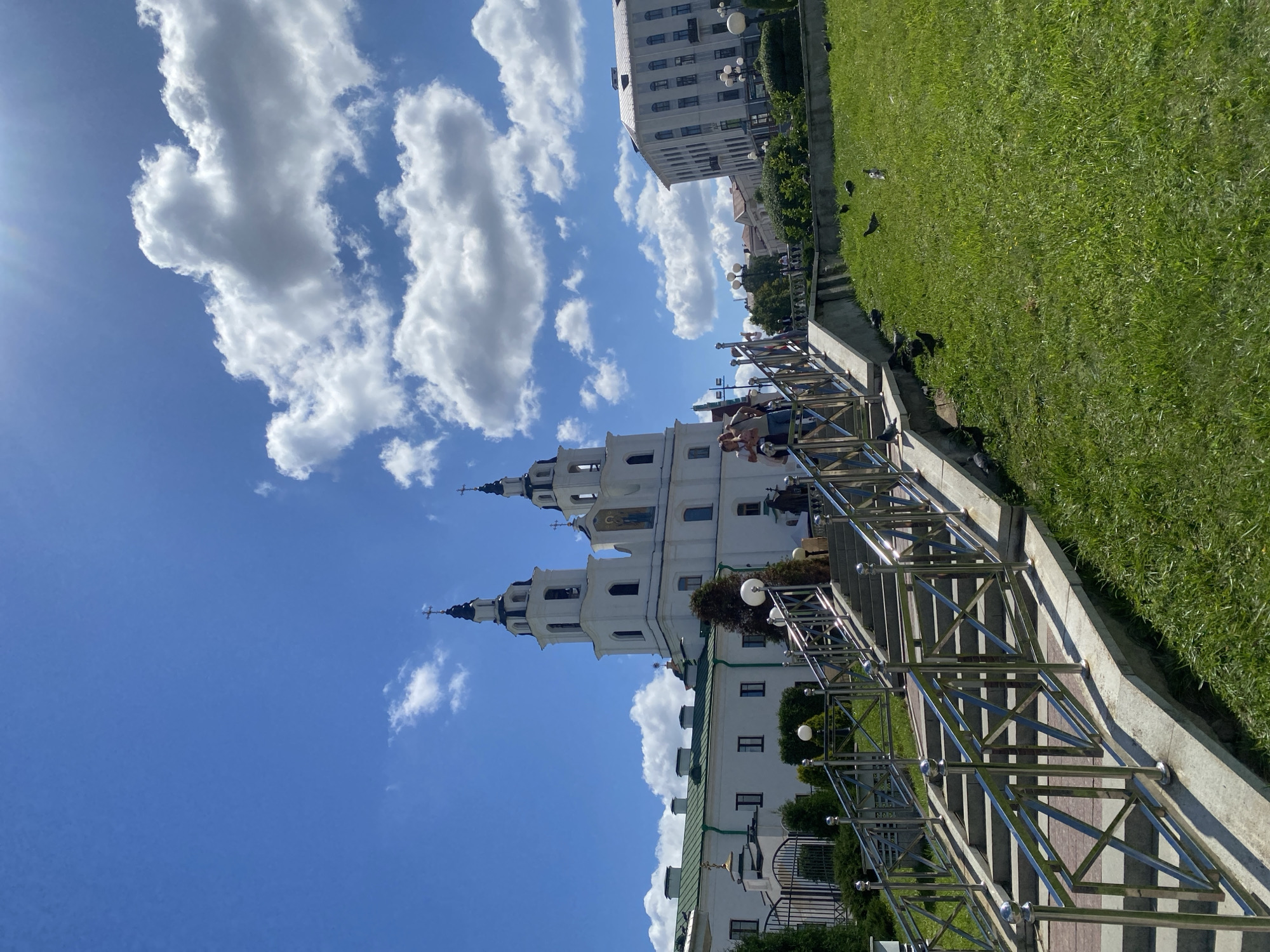 The image depicts a bright, sunny day in Minsk, Belarus, specifically at the Holy Spirit Cathedral. The cathedral is a prominent white building with two tall spires, each topped with a cross. In front of the cathedral, a wide, paved staircase with gleaming silver railings ascends. Several people are visible on the stairs, some ascending, others descending, engaging in casual movement. The staircase is flanked by lush green grass on the right, where several pigeons are scattered. To the left of the stairs, a fence encloses a well-maintained area with greenery. In the background, other buildings and trees contribute to the urban landscape, all under a vibrant blue sky dotted with fluffy white clouds. The sun is shining brightly, casting shadows and creating a slight lens flare in the upper right corner of the image.