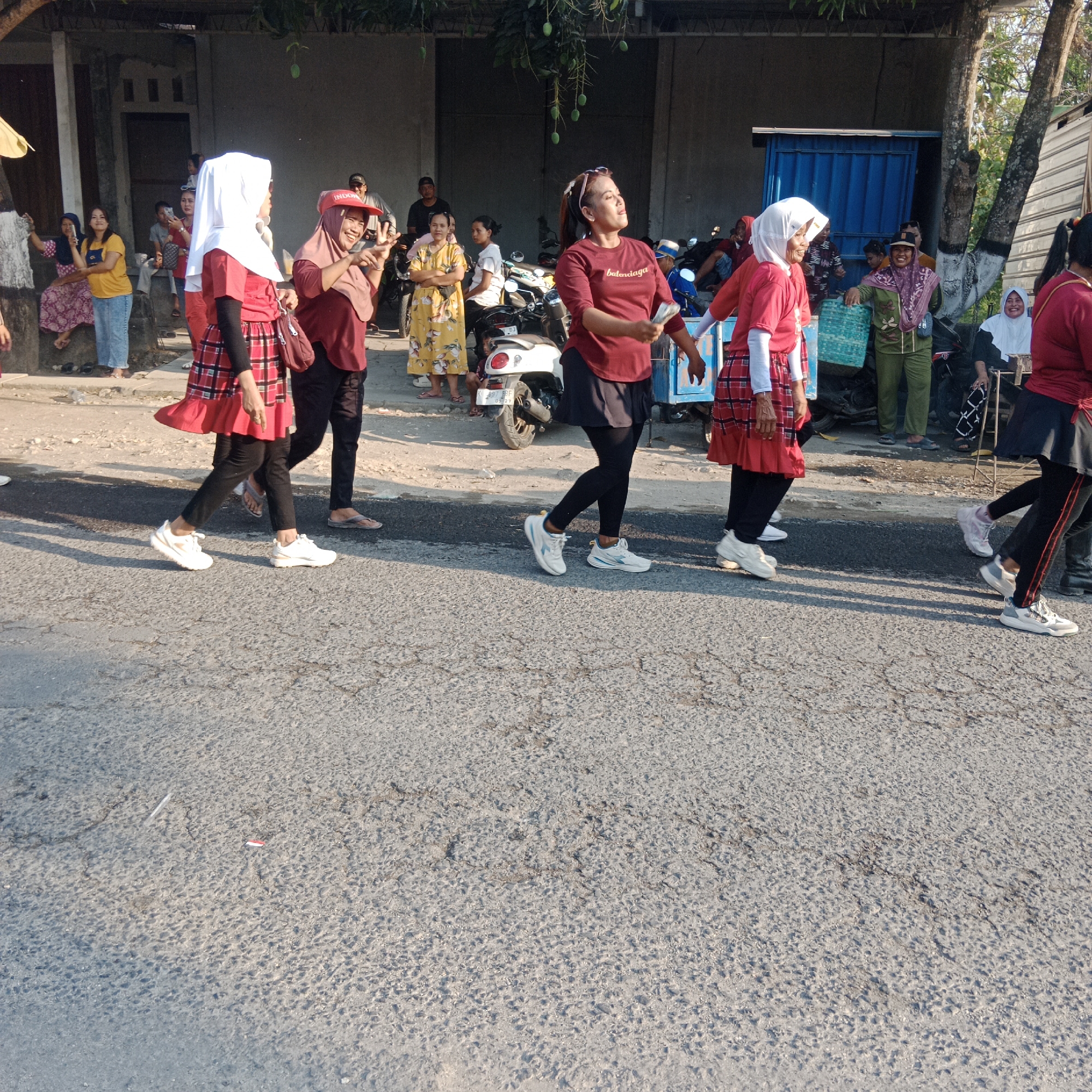 In the city of Tegalrejo, Indonesia, a group of women are walking down a street. They are wearing various colorful outfits, including hijabs and patterned skirts. Some of the women are smiling and looking towards the camera. In the background, there are buildings, a few motorcycles, and some onlookers. The scene appears to be a daytime event, possibly a parade or a community gathering. The road is paved and shows some signs of wear.