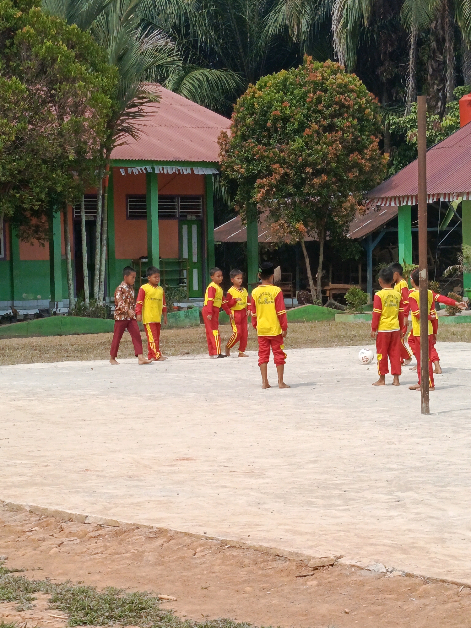 In a schoolyard in Megang Sakti IV, Indonesia, a group of young students in red and yellow uniforms are playing soccer. The ground is a dry, sandy surface, and in the background, there are two buildings with orange walls and red corrugated metal roofs, surrounded by lush green trees and palm fronds. A volleyball net pole stands on the right side of the frame. The students appear to be engaged in a friendly game, with some kicking the ball and others observing. The scene is brightly lit by sunlight, suggesting it is daytime.