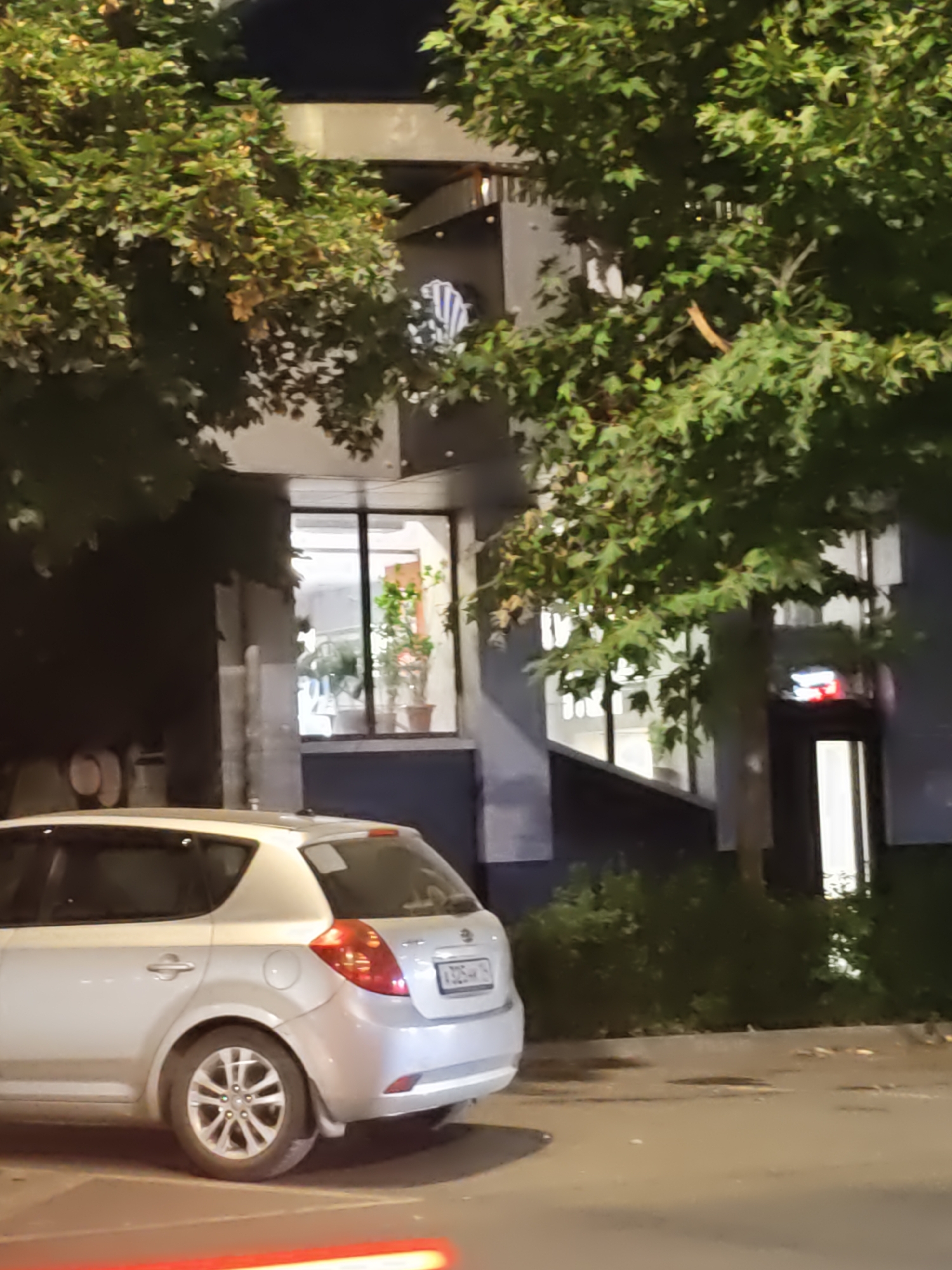 The image depicts a silver hatchback car parked on an asphalt surface. The car is positioned in the foreground, facing towards the right side of the frame. Behind the car, a building with large windows is visible, partially obscured by trees with green foliage. The building appears to be a multi-story structure. The scene is illuminated by artificial light, suggesting it is nighttime or a dimly lit environment. The license plate on the car is partially obscured but appears to have Cyrillic characters and numbers. The environment suggests an urban setting.