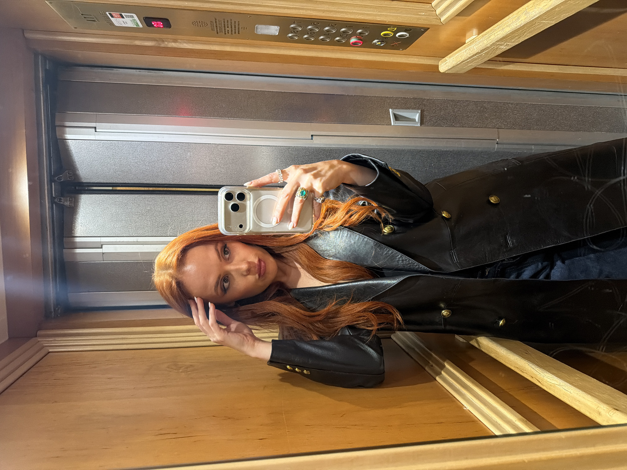 A woman with long, auburn hair is taking a selfie in an elevator. She is wearing a black leather jacket with gold buttons and multiple rings on her fingers. Her phone is a white iPhone with a clear case. The elevator has wooden paneling and a control panel with buttons labeled with floor numbers. The elevator appears to be ascending as indicated by the red floor indicator showing "00" and "01" above it. The location is likely Paris, France, given the design of the elevator.