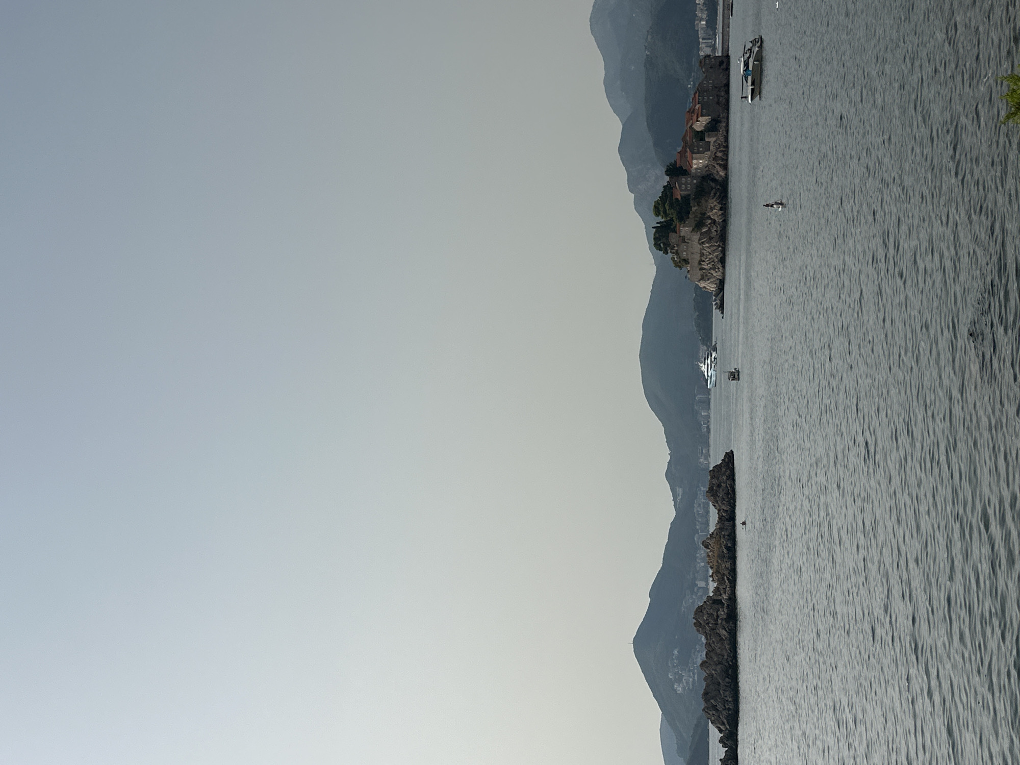 This is an eye-level, medium shot, outdoor photograph of Sveti Stefan, Montenegro, taken from a distance across the water. The scene depicts a clear, calm body of water in the foreground, with gentle ripples on its surface. In the middle ground, a small, rocky island is visible, adorned with red-tiled buildings and lush green trees. Several boats are scattered across the water. A white yacht is docked near the island, and a smaller white boat is moored to the right. A person is standing on a paddleboard in the water between the island and a rocky outcrop. Further in the distance, a row of mountains forms the backdrop, with a hazy, pale sky overhead. The overall atmosphere is serene and picturesque.