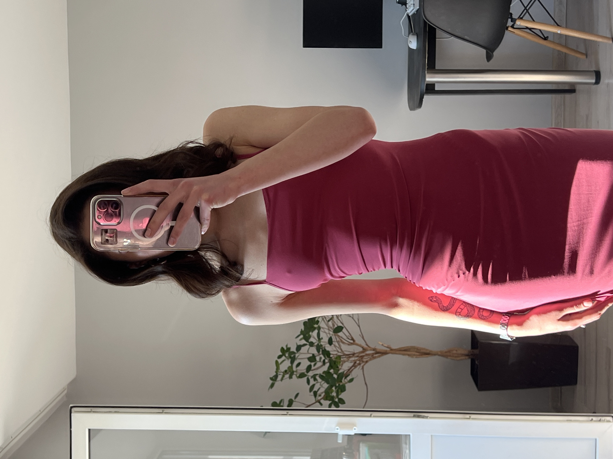 A young woman, with dark hair and fair skin, stands in a well-lit room, holding a pink smartphone to obscure her face. She is wearing a form-fitting, sleeveless, magenta dress. A tattoo of a snake is visible on her left arm. To her left, a tall, thin plant in a dark pot stands near a white doorframe. To her right, a black desk with a computer monitor, keyboard, and mouse is visible, along with a black chair with wooden legs. The room appears to be a bedroom or a studio apartment. The lighting suggests it is daytime. The image does not contain information that could place it in Ternopil, Ukraine.
