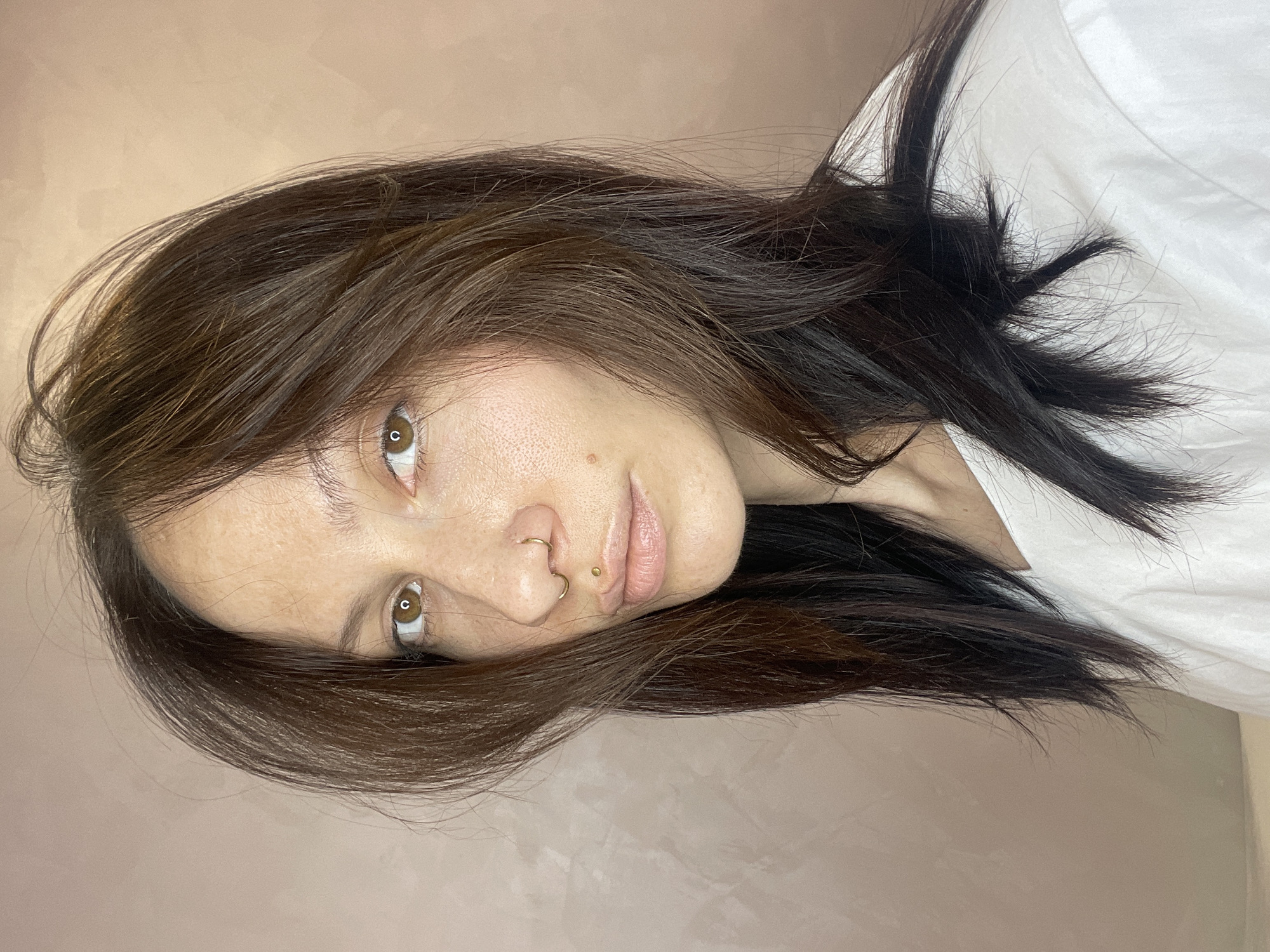 A close-up portrait shows a young woman with long, dark brown hair that falls across her shoulders and partially frames her face. Her head is tilted, and she gazes directly into the camera with warm brown eyes. Her complexion is visible, showing natural skin texture and some light freckles. She has two gold-toned facial piercings: a hoop in her septum and a stud above her upper lip. Her lips are slightly parted, revealing a natural, light pink hue. She wears a white garment, possibly a shirt or blouse, which is visible on her upper right side. The background is a plain, light beige or pinkish wall, indicating an indoor setting. The lighting is soft and even, creating a gentle and focused atmosphere. No specific activity is depicted other than posing for the portrait. The overall impression is one of calm engagement. The location of the photo is Izhevsk, Russia. No text is visible in the media.