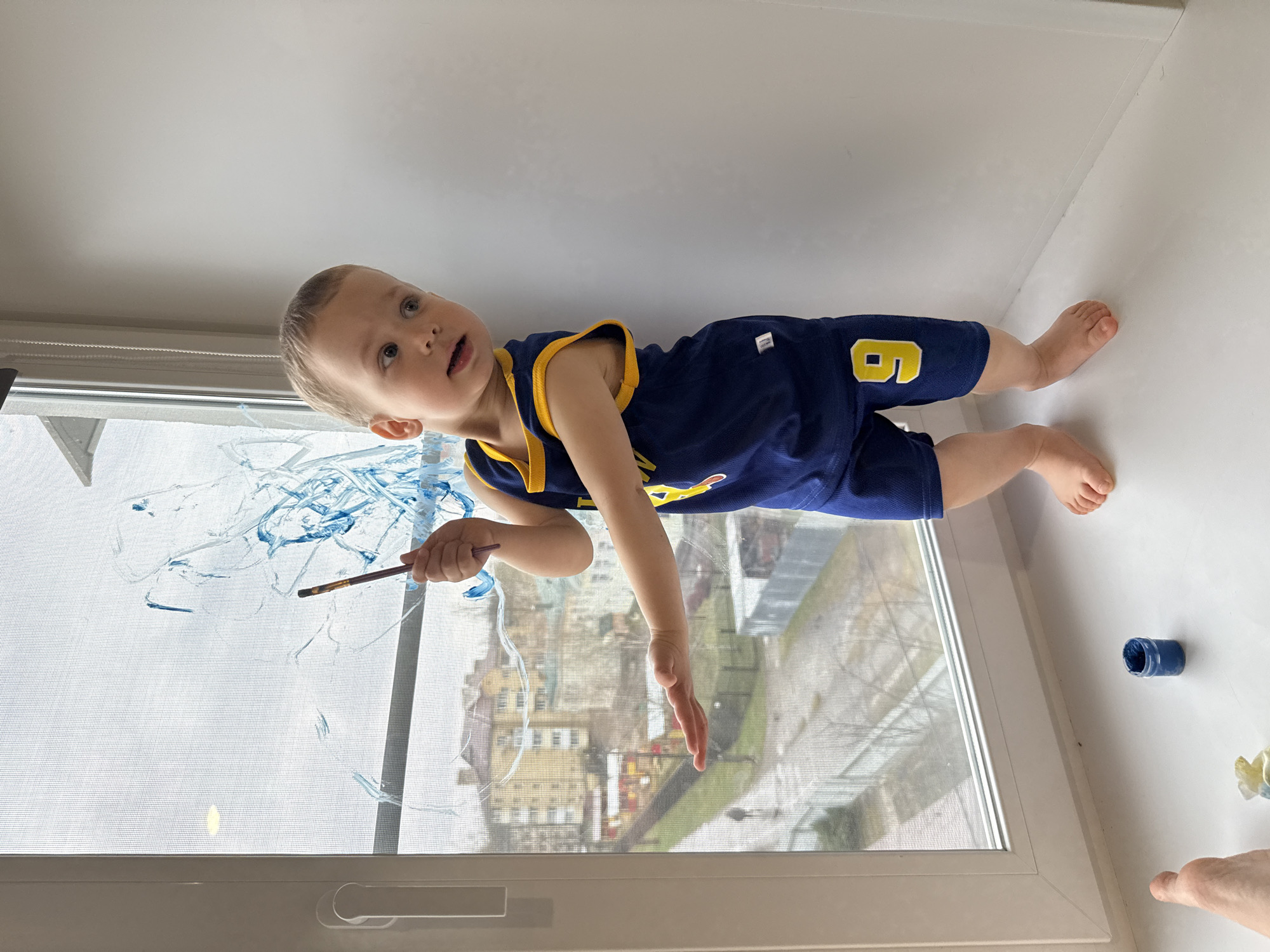 A young child wearing a blue and gold jersey with the number "9" is lying on a white windowsill, painting on a window screen with blue paint and a brush. A jar of blue paint is also on the windowsill. The view through the window shows buildings, which may be located in the city of Rusanovo, Russia.