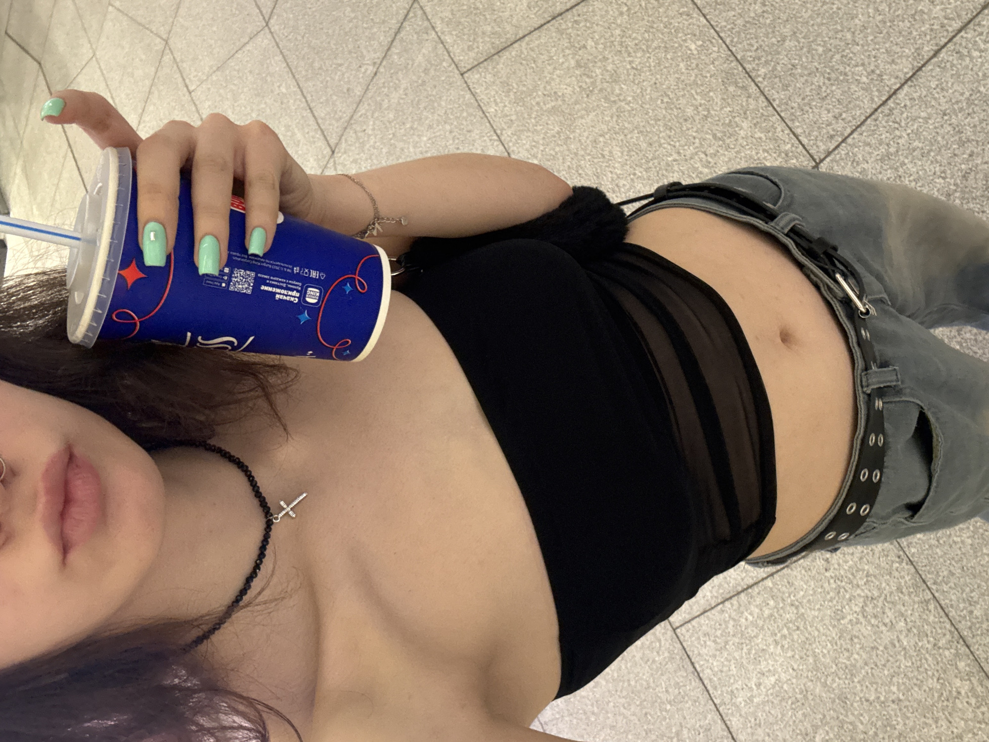 Here is a detailed description of the image:
**People:** The image shows a young woman holding a Burger King cup with a straw. Her nails are painted a light green color. She is wearing a black tube top and jeans with a belt. She also has a necklace with a cross pendant and a nose ring.
**Objects:** The primary objects are the Burger King cup, the woman's clothing, the necklace, and the nose ring. Other noticeable objects include the straw in the cup and the belt on her jeans.
**Actions/Interactions:** The main action is the woman holding the cup. She is presumably taking a photo of herself.
**Environment:** The background appears to be a tiled floor. It is a public area, possibly a shopping mall or transit station. Based on the cup details and QR code details, the cup is in Russian, so the country is more than likely to be in Russia. Based on the QR code information, Saint Petersburg, Russia may be an option.