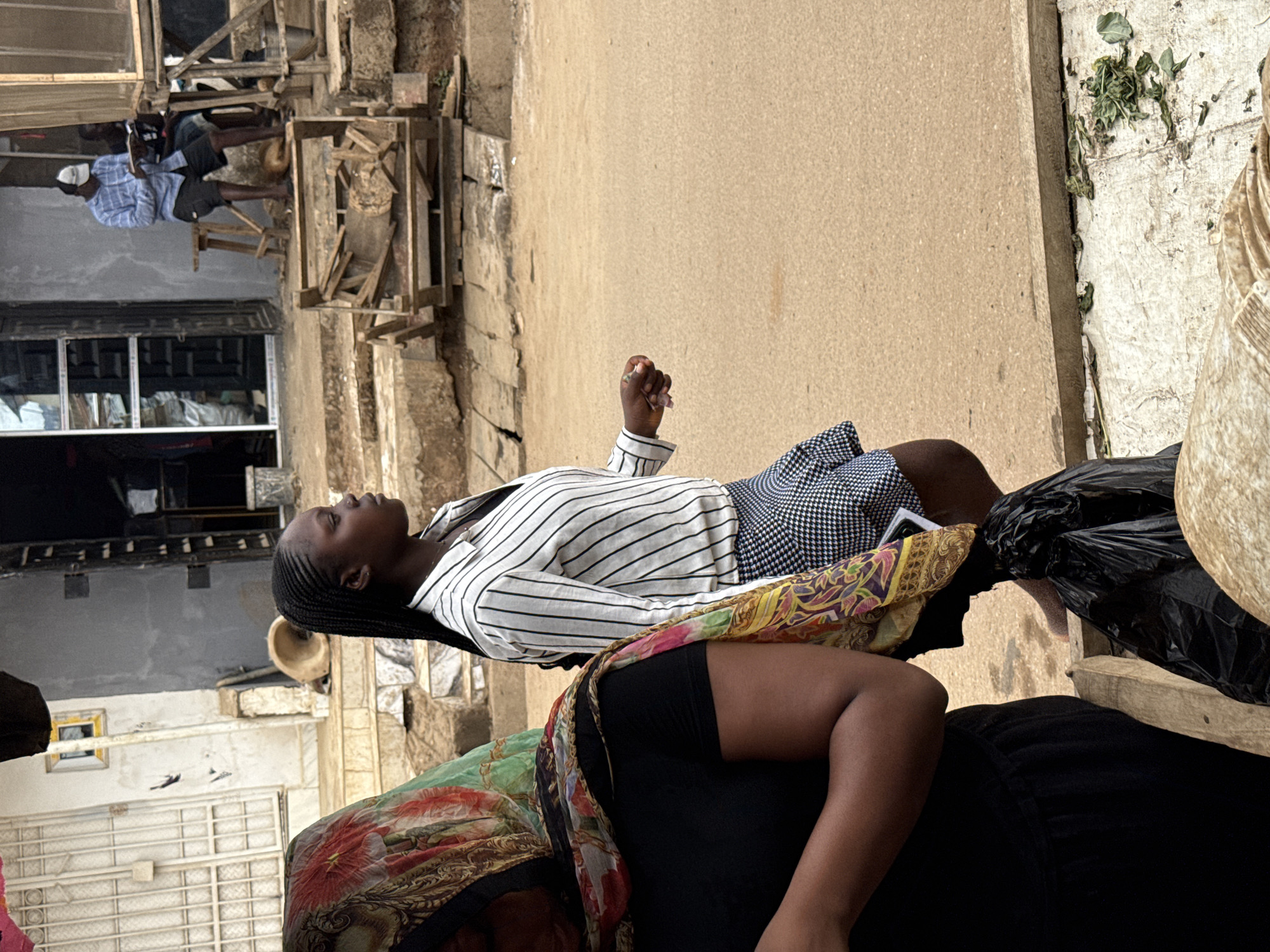 A full shot shows a Black woman with braided hair lying on what appears to be a black cushion or seat covered in a colorful scarf or patterned fabric, potentially taking a nap. She is wearing a horizontally striped white and black top, and a short, blue patterned skirt. Her arm is raised slightly, with her hand partially clenched. The background is a bustling street scene. In the upper-left of the frame, a person is seated and wearing a face mask and a blue plaid shirt. There is a structure with wooden beams in front of a building with windows behind them. The buildings are weathered and show signs of age. The ground and walls are light brown, and there are some plants or greenery growing along the wall in the upper-right of the frame. Based on the overall style and architecture, and the skin tones of the individuals, the location may be Abuja, Nigeria.