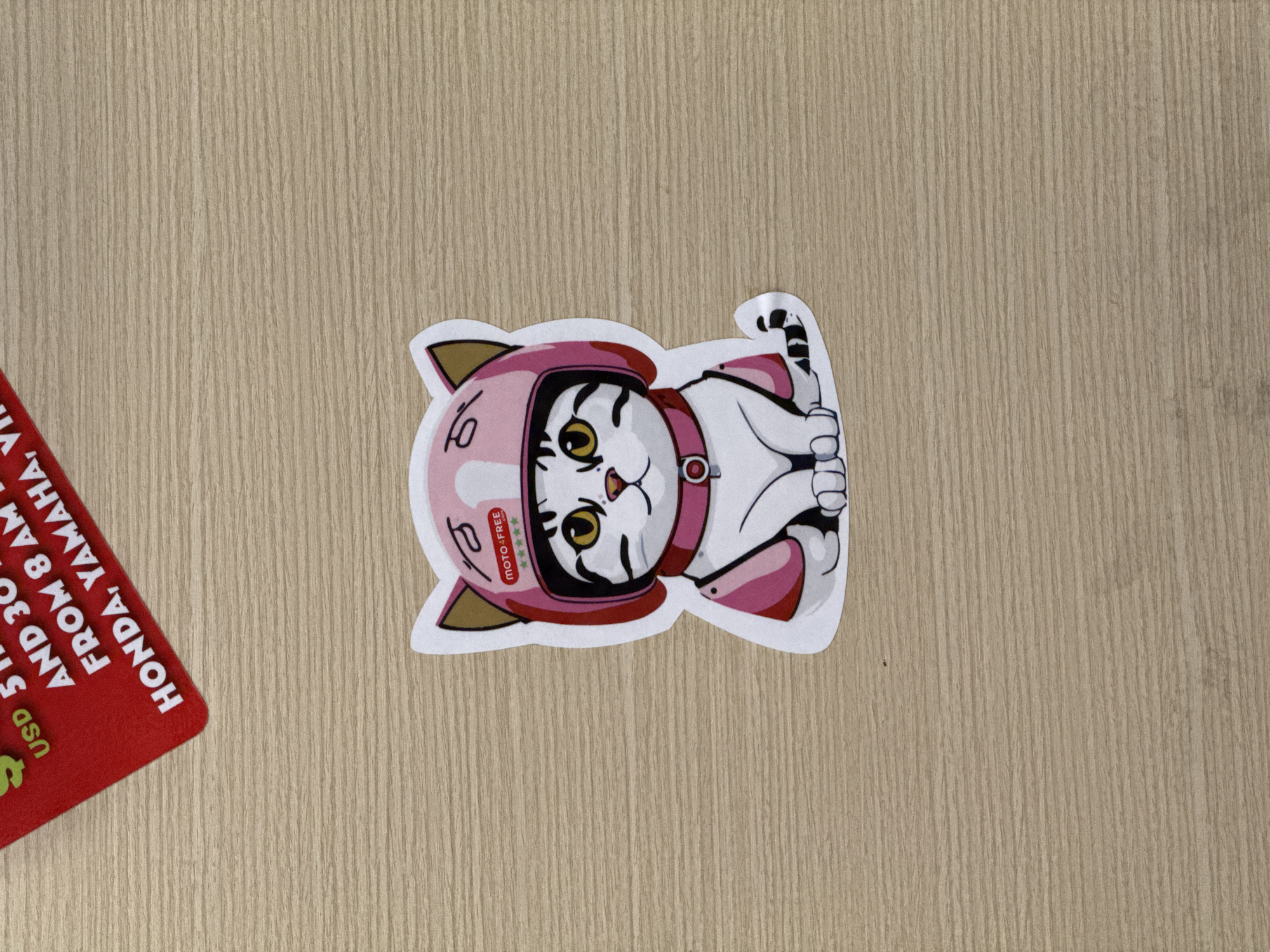 The image displays a playful cat sticker and a partial red card resting on a light brown wooden surface in Nha Trang, Vietnam. The scene is shot from directly above, highlighting the textures of the objects and background.
The main subject is a white cat sticker featuring black stripes and striking yellow eyes. The cat wears a vibrant pink helmet, complete with small cat ears, and a matching pink collar or harness. A small pink tongue is visible, adding to its cute expression. On the helmet, there's a small logo that reads "MOTO-FREE" with five accompanying stars, and another stylized symbol resembling "lo". The sticker has a clean white outline, contrasting with the background.
In the bottom-left corner of the frame, a segment of a red card is visible. It features white capitalized text, partially obscured. Readable phrases include "USD", "AND 30", "FROM 8 AM", and "HONDA, YAMAHA, VI". The wooden surface exhibits a distinct vertical grain pattern, suggesting it is either light-colored wood or a laminate. The overall lighting is soft and even, indicating a well-lit indoor setting. No people are present in the image.
