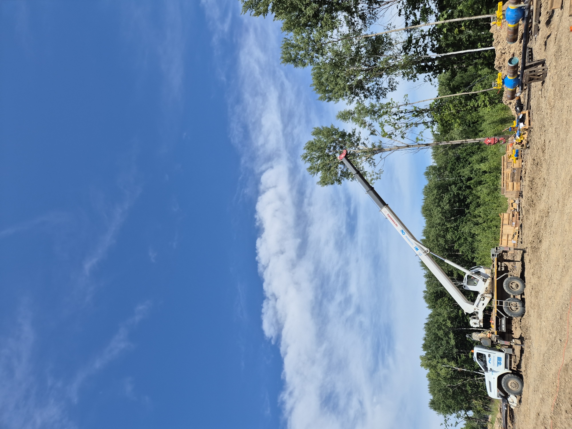 In Taezhnoe, Russia, on a clear day with a blue sky and scattered clouds, a construction scene unfolds. A white crane truck is positioned on uneven, sandy terrain, its boom extended upwards and diagonally to the right. The truck has a white cab with blue decals and appears to be a utility vehicle. Various construction materials, including wooden crates and large blue and yellow valves connected to pipes, are scattered around the site. A dense forest of green trees forms the backdrop, with some taller trees standing out in the foreground. The ground is a mix of dirt and sand, with some orange marking lines visible. The overall impression is a construction project in progress in a rural, wooded area.