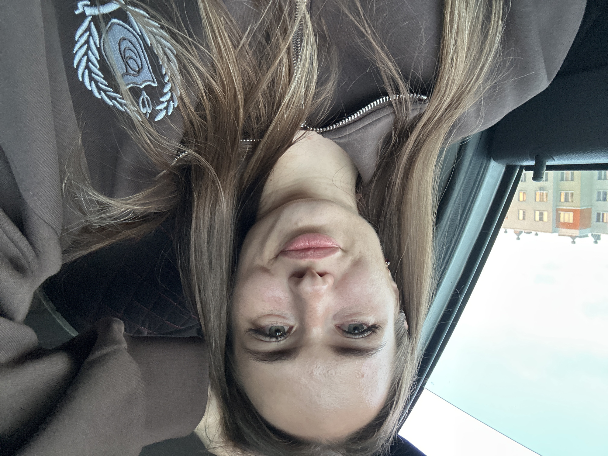A woman with light brown hair, wearing a brown zip-up hoodie with an embroidered crest on the right side, is pictured from the chest up. She is inside a vehicle, with a window on the left showing buildings in the background. The woman is looking directly at the camera and smiling faintly. The lighting suggests it may be daytime.