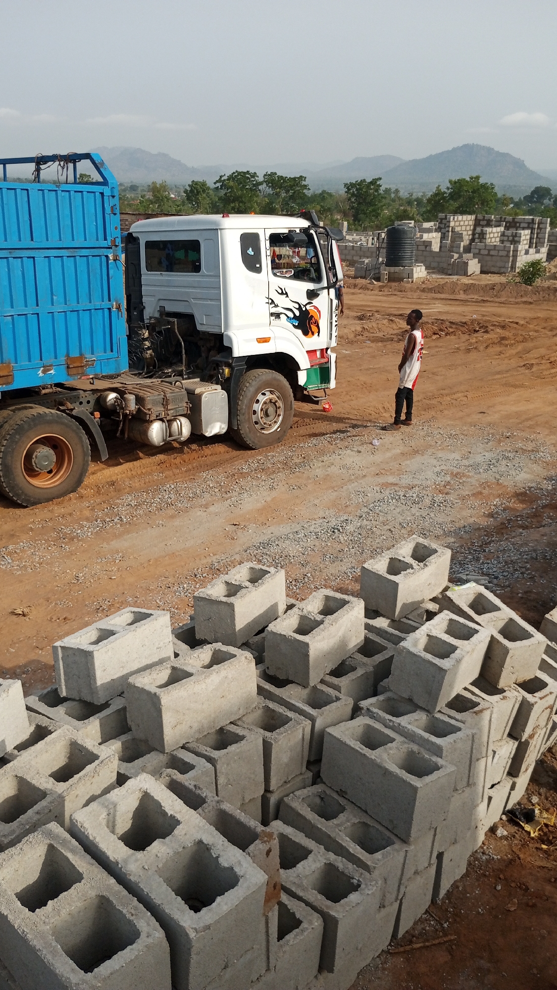 The image shows a construction site in a location identified as Unknown, Unknown. In the foreground, a stack of grey concrete blocks fills the lower portion of the frame. Beyond the blocks, a blue and white truck is parked on a dirt surface covered with gravel. Adjacent to the truck is a young man in a white and red jersey and black pants. In the background, there is a building under construction, a black water tank, and green trees. In the far distance, hazy mountains can be seen.