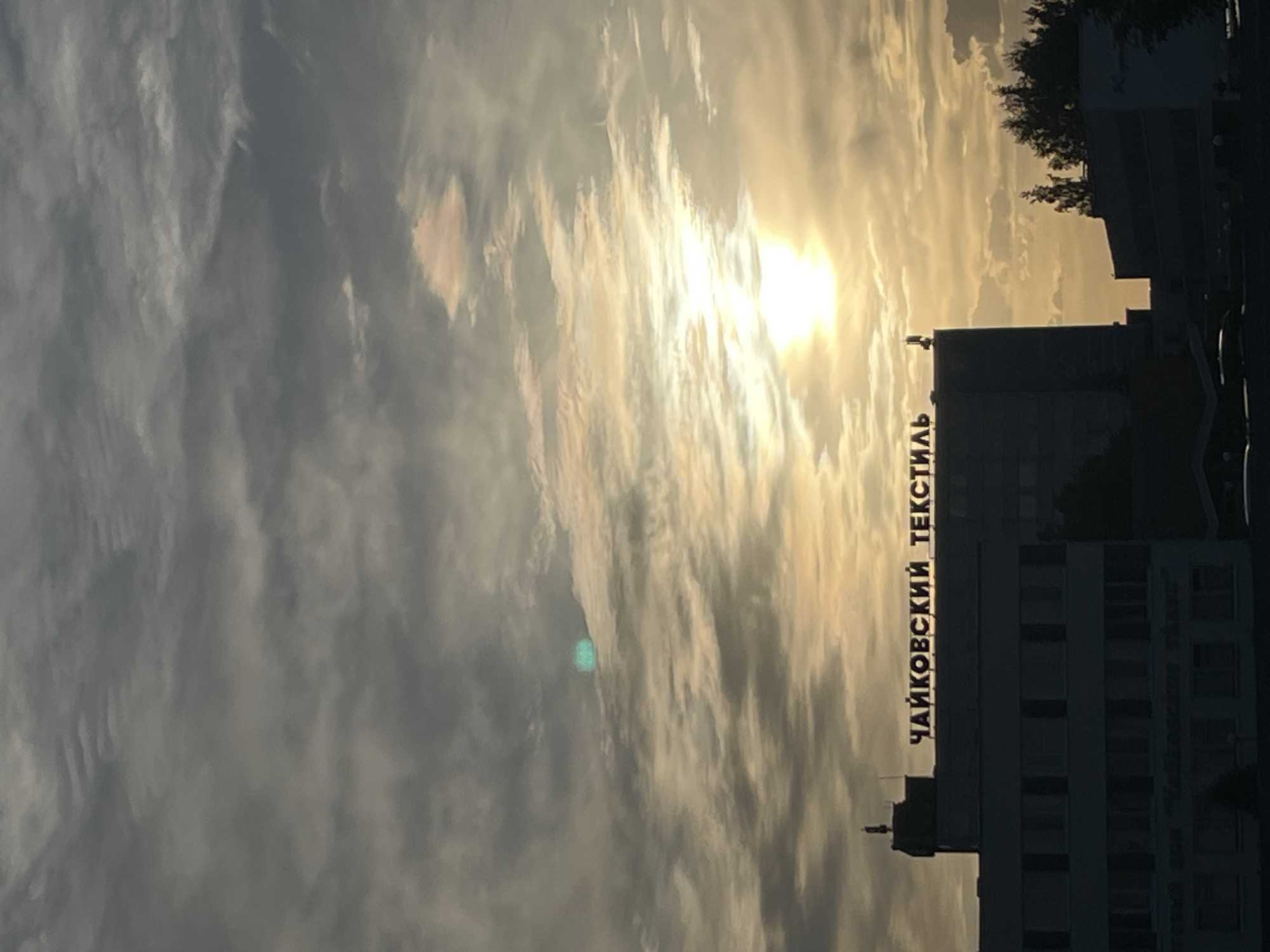 The image shows a building with the text "ЧАЙКОВСКИЙ ТЕКСТИЛЬ" (Chaikovsky Textile) on it, likely located in the city of Chaikovsky, Russia. The sun is setting or rising behind the building, creating a hazy and bright sky. The building is silhouetted against the sky, and some trees are visible at the edge.