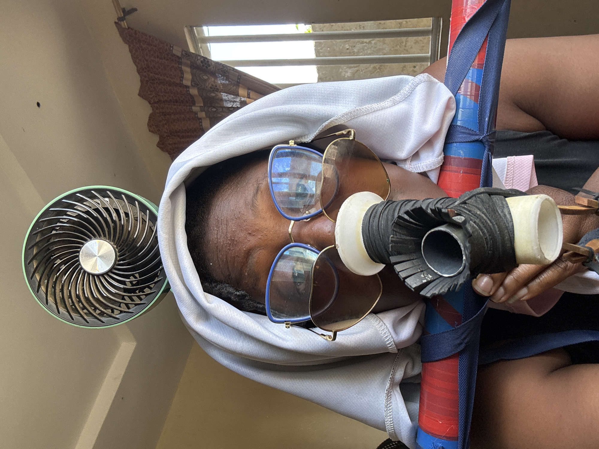 A close-up, waist-up shot captures a person wearing a white headscarf and sunglasses. They are holding a blue and red striped object wrapped in dark tape and a black object with a white nozzle, which is positioned in front of their mouth. A small, round fan with a green rim is visible above their head. The background includes a beige wall, brown patterned curtains, and a window with vertical bars. The image appears to be taken indoors in Jawu, Nigeria.