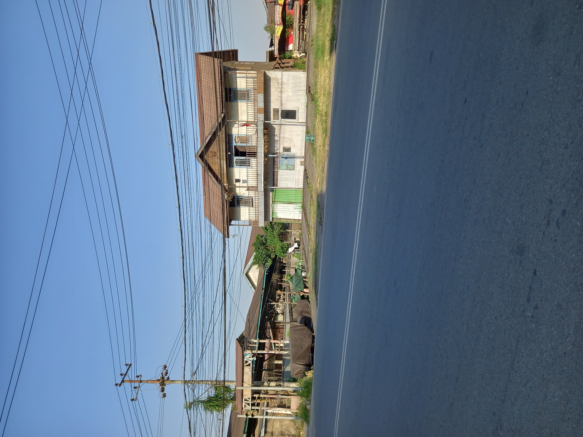 A sunlit outdoor scene depicts a two-story wooden building with a brown tiled roof, located on the side of a paved road. The building has a balcony on the upper level with a wooden railing and several windows. The lower level has a green door on the left, a blue-paned window, and a plain window. To the left of the main building, there is a covered structure with a brown roof, and several vehicles covered with brown tarpaulins. A wooden utility pole with numerous electrical wires stretches across the scene. The road in the foreground has white lane markings. In the background, a clear blue sky is visible, with more electrical wires crisscrossing. The building appears to be a shop or residence along a commercial or residential street in Samarinda, Indonesia.