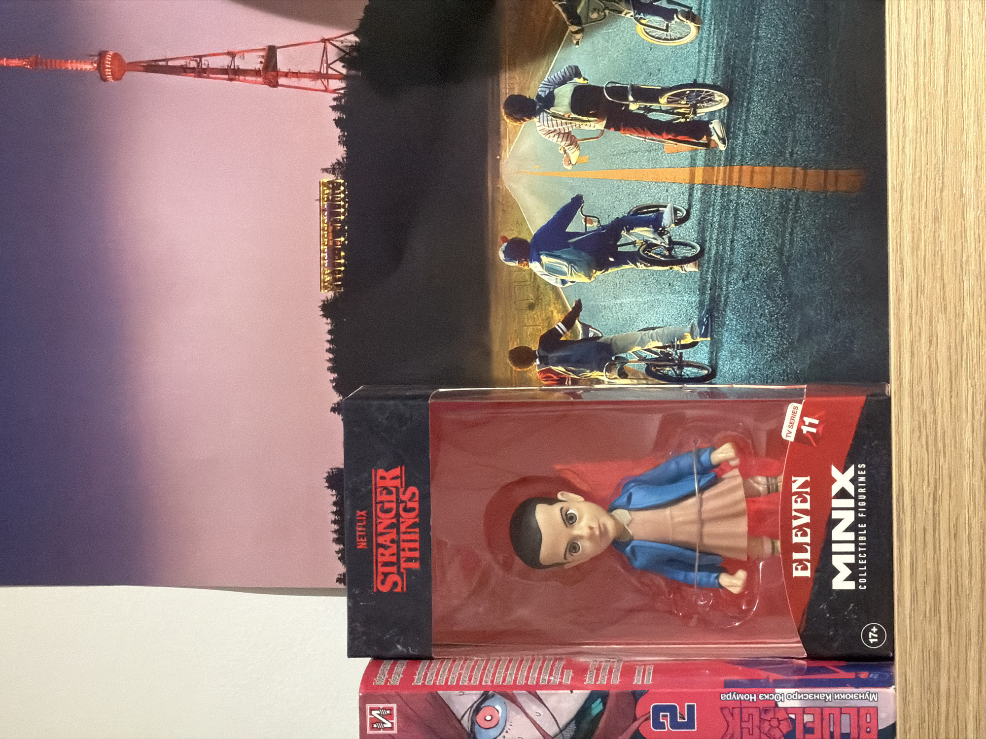 The image shows three objects: A pink and purple picture of the Tbilisi TV Broadcasting Tower, a picture of children riding bikes, and two unopened boxes: a 'Stranger Things' Eleven Minix collectible figurine and a blue-lock volume two manga.