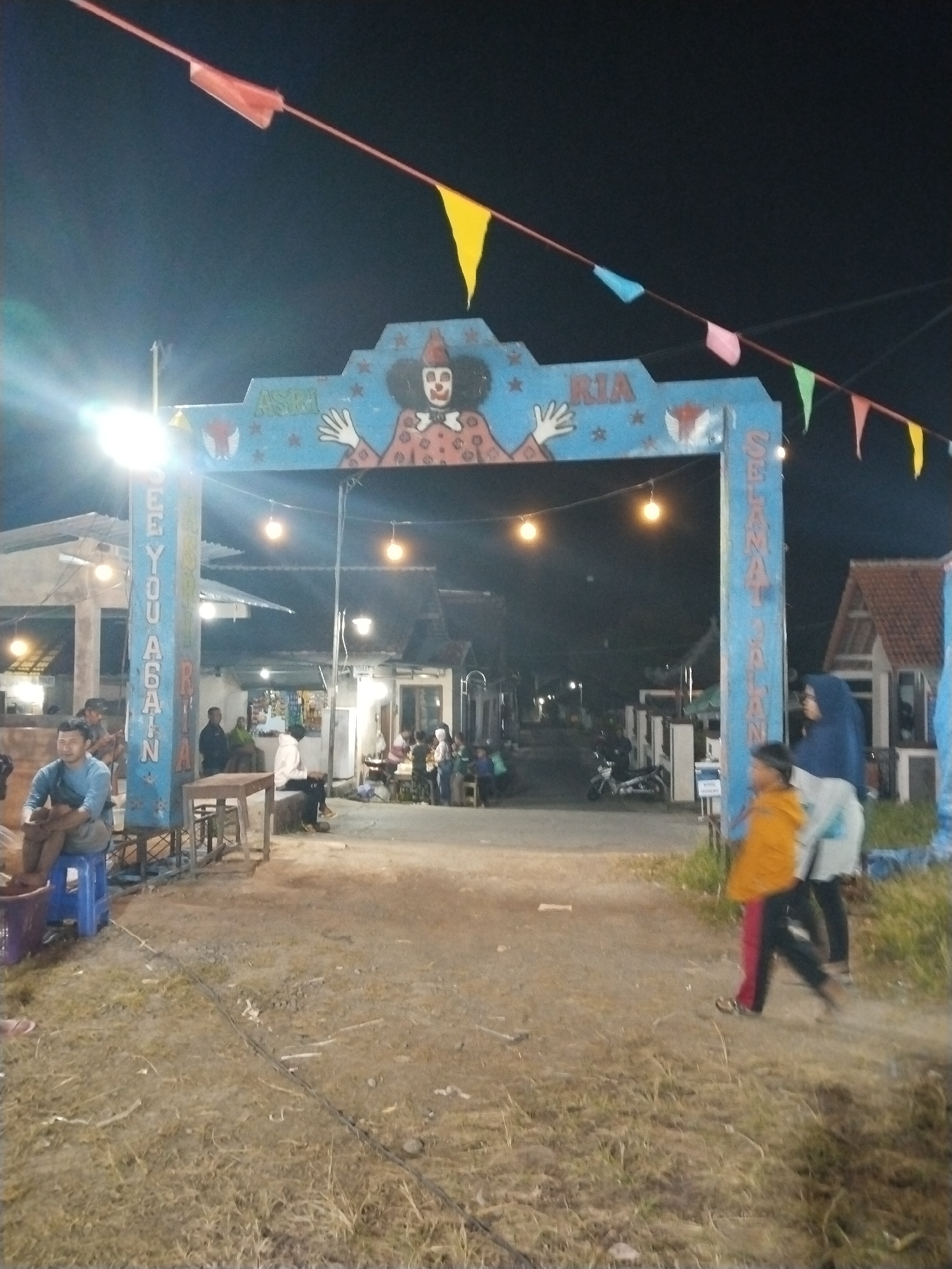 The scene is a nighttime view of a street in Seloromo, Indonesia, marked by a large, decorated archway spanning the road. The archway is painted blue and features a depiction of a clown-like figure at its center, flanked by the phrases "ASIH RIA" and "SEE YOU AGAIN," along with "SELAMAT JALAN" on its supports. Strings of colorful triangular flags are strung above the arch. Under the archway and beyond, the street stretches into the distance, lined with simple buildings and some individuals seated at tables, possibly at an outdoor cafe or gathering place. To the right of the arch, two figures are seen walking, one wearing a blue hijab. To the left, a man sits on a blue plastic stool, cradling a child or infant. The ground beneath the archway is mostly bare dirt or gravel. Overhead lighting is visible from the arch and the buildings, illuminating the scene.