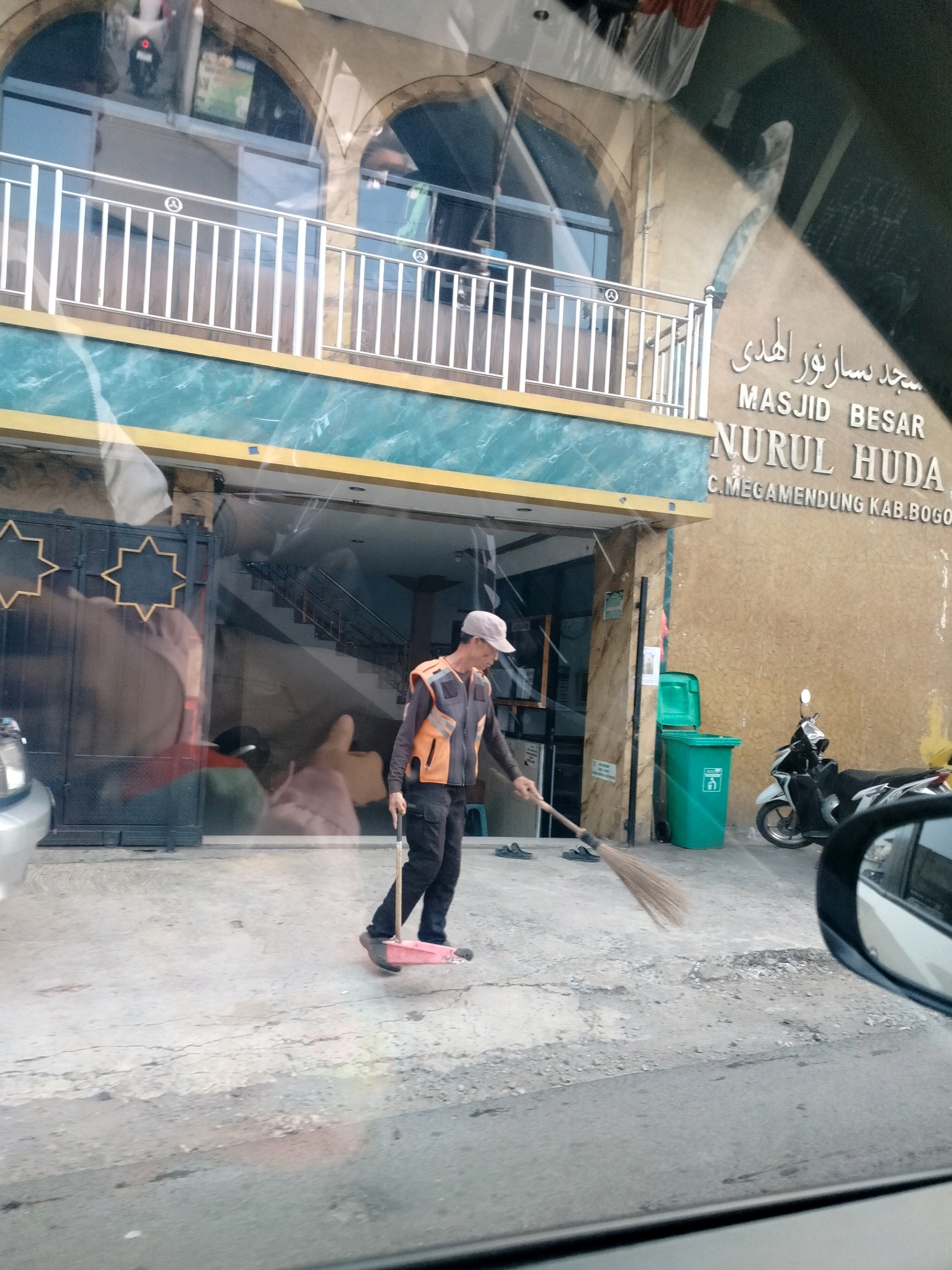 The image shows the facade of "MASJID BESAR NURUL HUDA" in C. Megamendung, Kab. Bogor, Indonesia. A man in a reflective orange vest and dark pants is sweeping the concrete ground with a broom while standing on a small red platform with wheels. A green trash can and a motorcycle are visible near the entrance of the mosque. The mosque has a cream-colored exterior with arched windows and a white railing on the upper level. The scene appears to be captured from a vehicle, as indicated by reflections on the glass and a side-view mirror in the foreground.