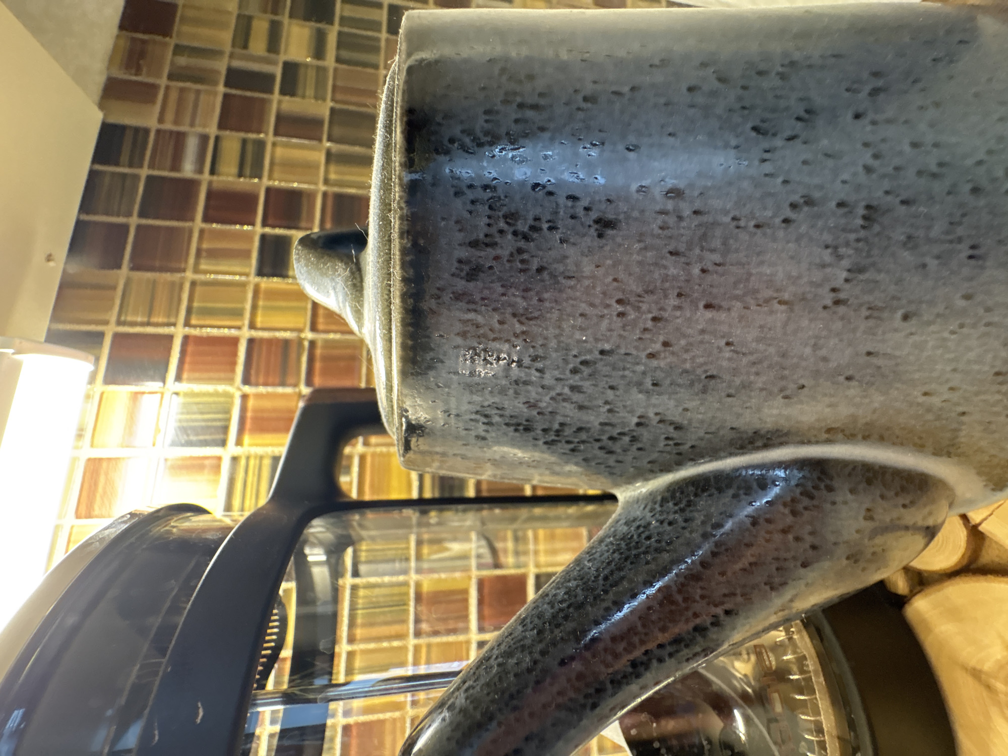A close-up shot depicts a grey ceramic mug with a textured surface, positioned near a black appliance, possibly an air fryer, against a backdrop of a mosaic tiled wall. The location, Chaikovsky, Russia, cannot be verified from the visual information provided.