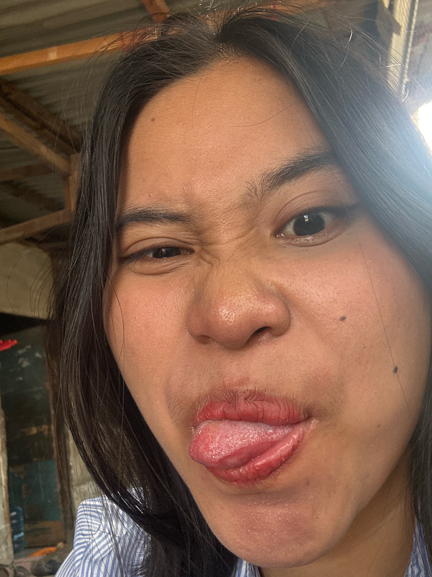The image is a close-up, outdoor selfie of a young woman with dark hair. She is winking with her right eye and sticking her tongue out. The background is out of focus and appears to be a rustic outdoor setting with a corrugated metal roof and wooden beams. The lighting suggests it is daytime. The woman is wearing a blue and white striped shirt. There are no discernible landmarks or specific details that would allow for precise location identification within East Jakarta, Indonesia.