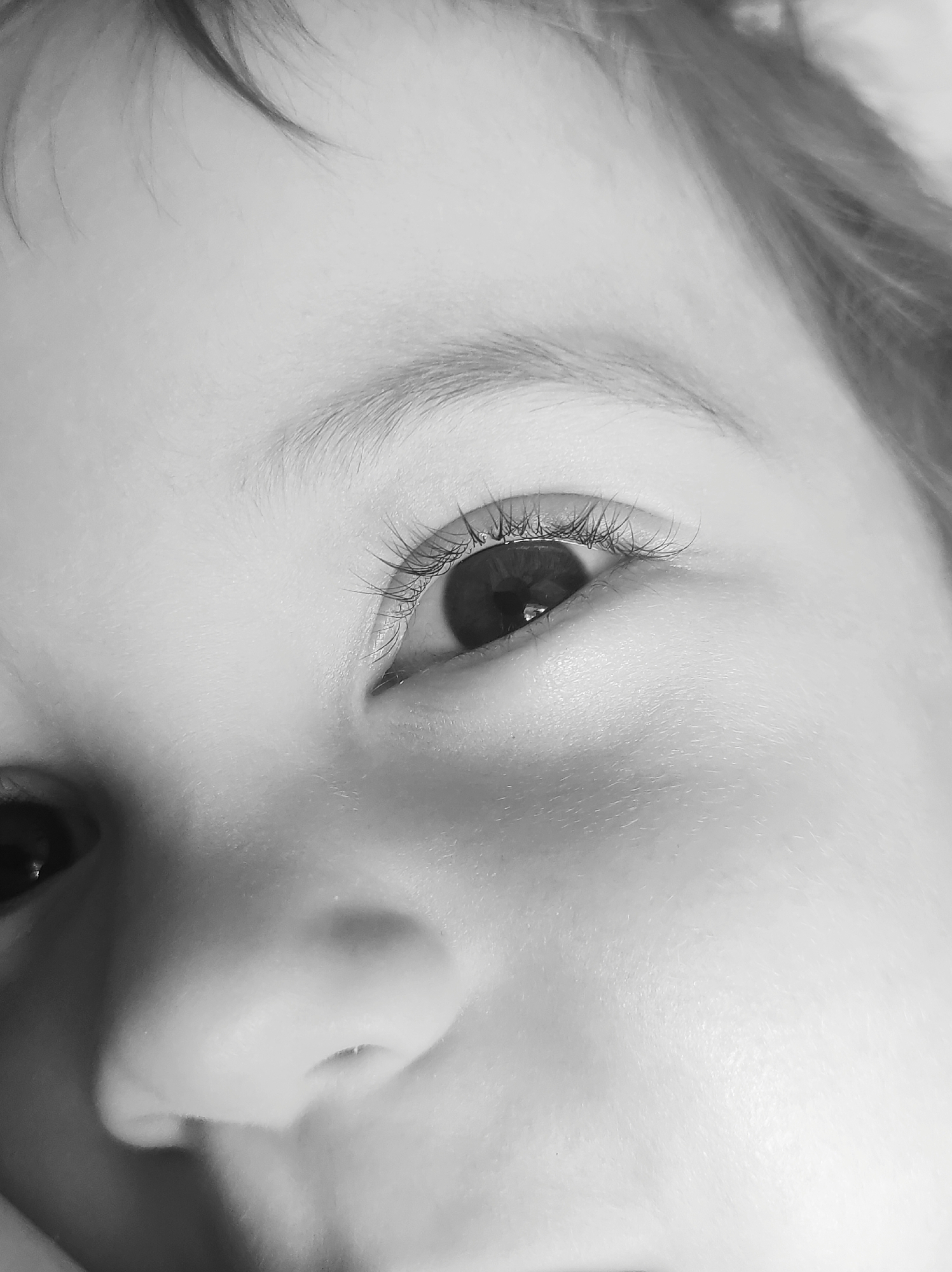 This is a close-up, black and white photograph of a baby's face, with a focus on the right eye and the bridge of the nose. The baby's skin is smooth, and fine baby hairs are visible near the forehead. The right eye is open, revealing a dark iris with a slight reflection. The eyelashes are prominent and delicate. The nose is small and rounded, with nostrils visible. The overall impression is one of innocence and youth. The photograph does not contain any information or context that would allow for its localization in the city of Chaikovsky, Russia.