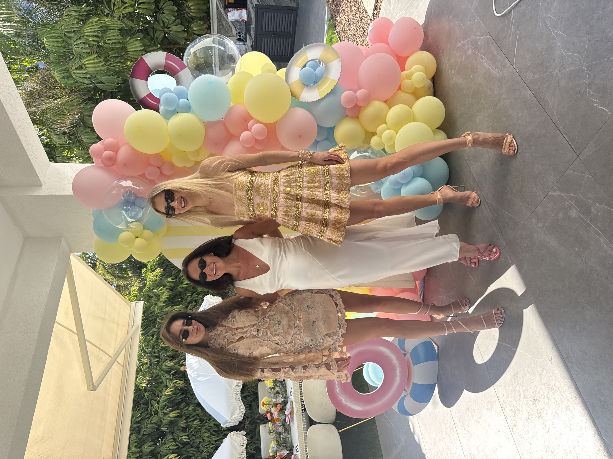 Three women are posing in front of a balloon arch in Agios Tychona Community, Cyprus. The woman on the left wears a beige lace dress and sunglasses, with her hair down. The woman in the middle is wearing a white satin dress and sunglasses, with her hair down. The woman on the right wears a sequined gold dress and sunglasses, with her hair down. Behind them, a balloon arch is decorated with pastel-colored balloons and a life preserver. In the foreground, there are inflatable pool rings. The ground is a tiled patio. In the background, there is a table set for a party with flowers, umbrellas, and chairs. The scene appears to be outdoors, possibly at a backyard party or event.