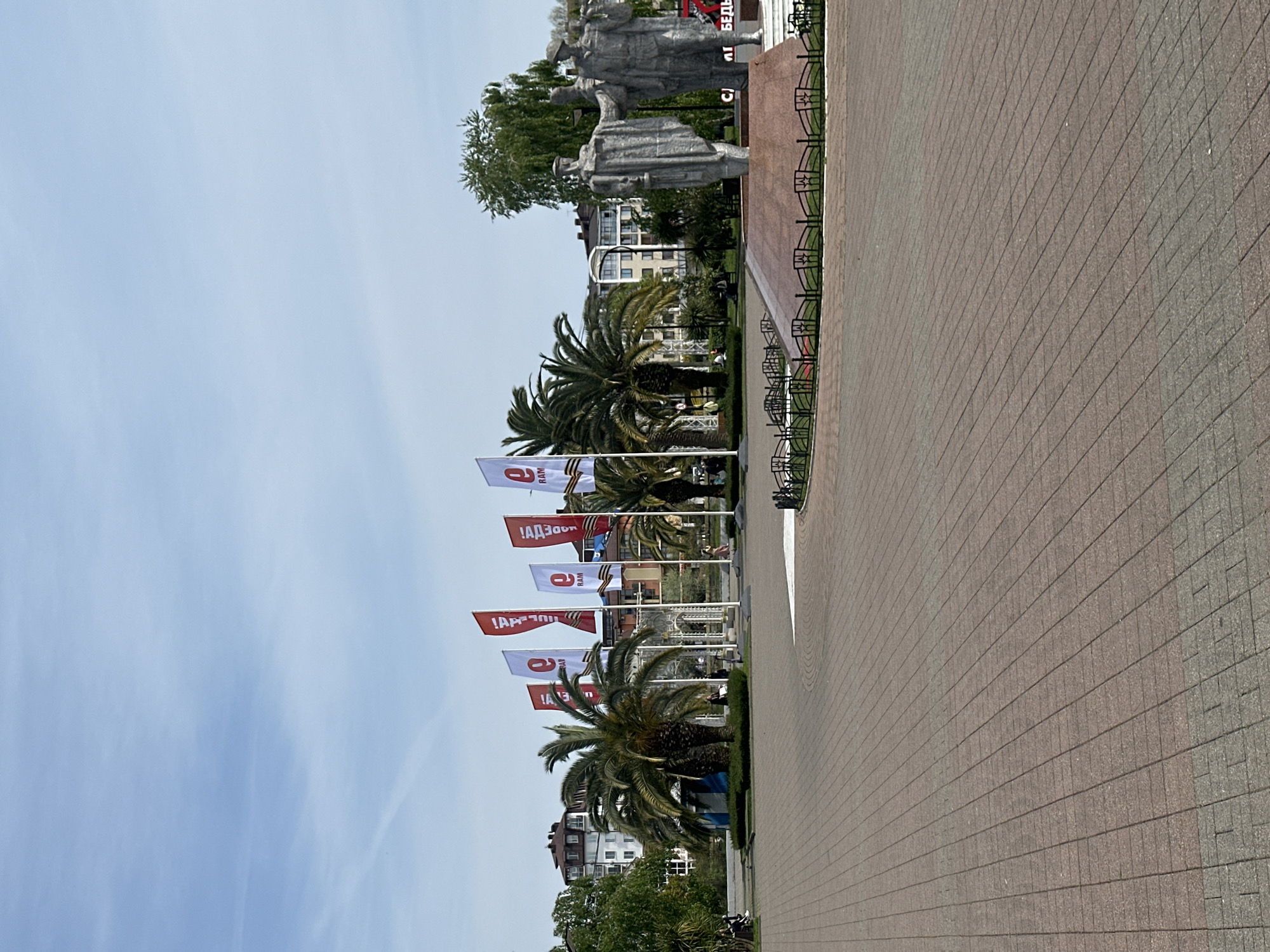 The image captures an outdoor public square or promenade in Sochi, Russia, on a clear, bright day. The scene is dominated by a large, grey stone monument depicting several stern-faced figures, likely soldiers or military personnel, standing upright. The monument rests on a reddish-brown base.
In the mid-ground, a row of flagpoles displays white and red flags. The white flags clearly feature the text "9 МАЯ" (9 MAY) along with a representation of the St. George ribbon, a symbol associated with Victory Day in Russia. The red flags also bear Cyrillic text, partially visible, which includes phrases like "ДА ЗДРАВСТВУЕТ" (Long Live) and likely refers to Victory Day celebrations, complementing the "9 МАЯ" theme. Several lush palm trees are visible throughout the area, alongside other green foliage and trees, indicative of a warm climate.
In the background, behind the monument and flags, multiple-story buildings with light-colored facades are visible. The foreground consists of a broad, paved area made of brown and grey interlocking bricks or pavers, with a decorative black metal fence running along its left edge. No distinct people are visible, but the setting suggests a public space for commemoration or gatherings. The time of day appears to be daytime with clear skies, and the overall impression is one of solemn remembrance and national pride, likely related to the annual Victory Day celebrations.