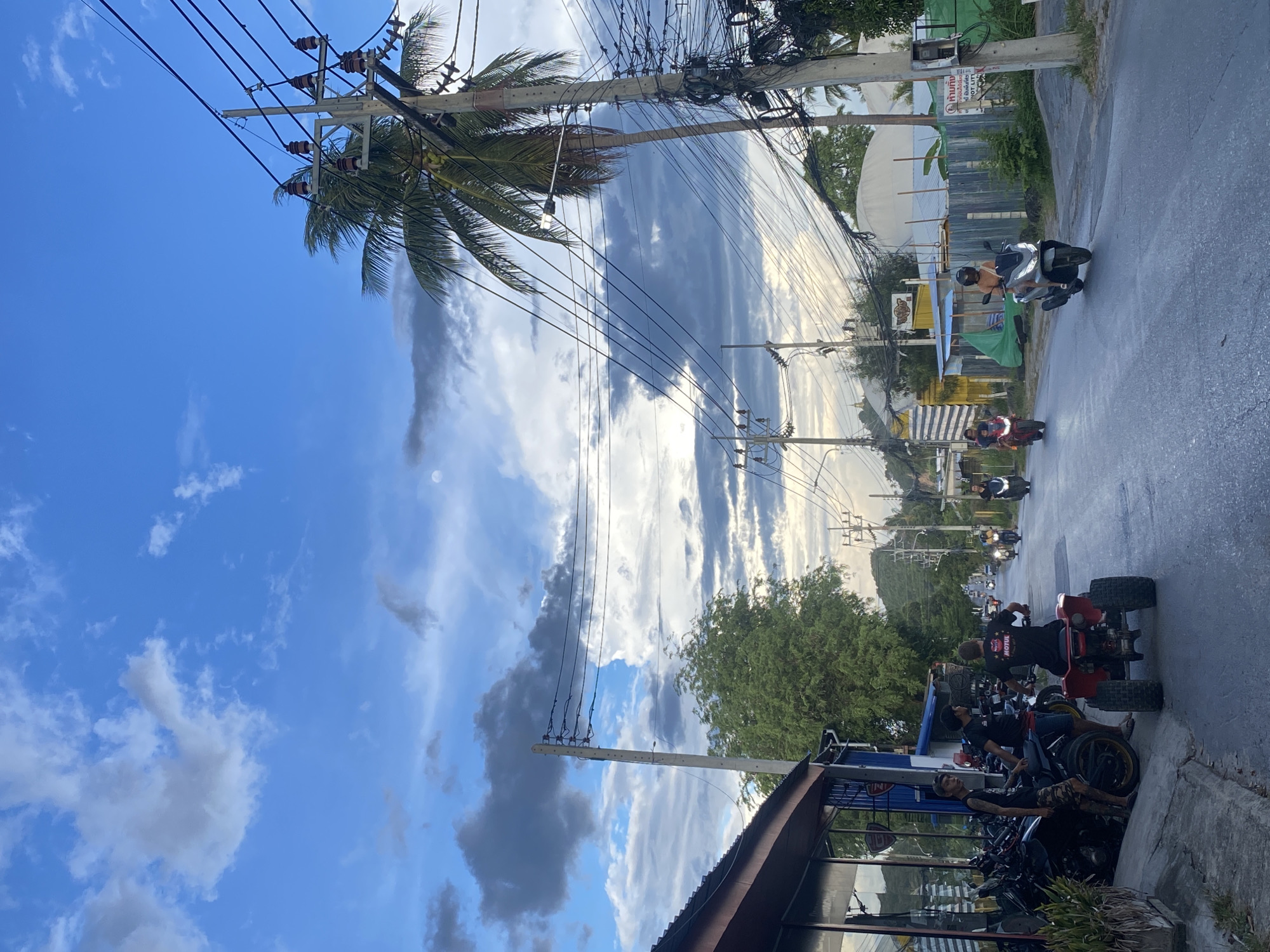 The image depicts a street scene in บ้านบางรักษ์, Thailand. The sky is predominantly blue with scattered cumulus clouds. Power lines and utility poles are visible throughout the scene. A palm tree is present, extending from one of the utility poles.
A street runs through the middle of the frame. Several individuals on scooters are moving along the street. Other people and several parked motorcycles are visible on the right side of the street. A red ATV is visible among the parked motorcycles. Buildings are visible on both sides of the street. The street appears to be wet or recently washed.