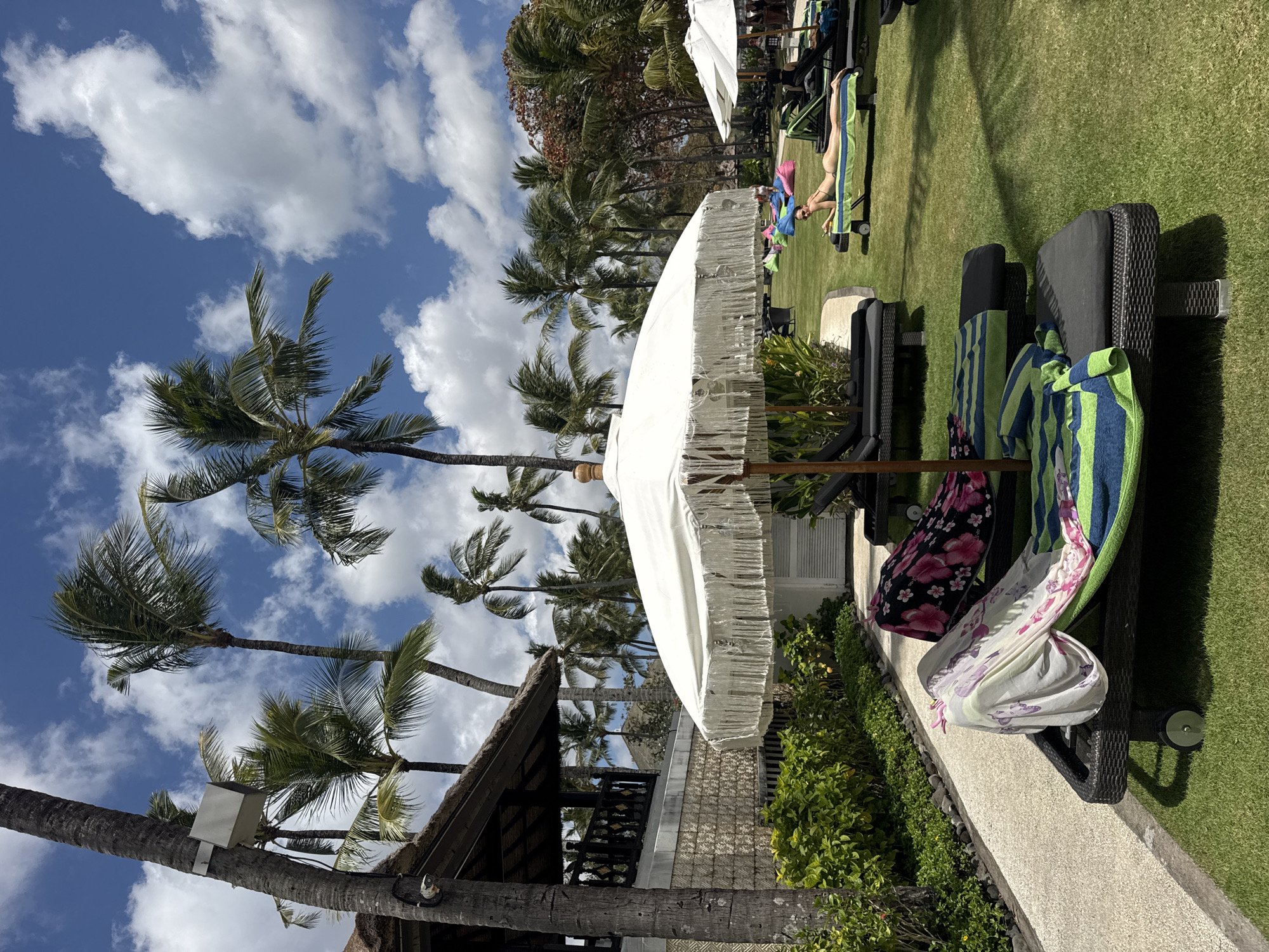 A sunny day in Kuta Selatan, Indonesia, features a resort's outdoor area with lush green grass, palm trees, and lounge chairs. In the foreground, a white fringed umbrella shades two unoccupied lounge chairs, each covered with towels, one with a floral pattern and the other with striped blue and green towels. Further back, several other lounge chairs are visible, some occupied. A woman in a bikini is sunbathing on a lounge chair in the middle ground. In the background, more lounge chairs, beanbag chairs in various colors, and other resort structures are scattered across the lawn. The sky is a vibrant blue with scattered white clouds. A pathway runs along the left side of the image, bordered by dense green bushes and a building with a thatched roof.