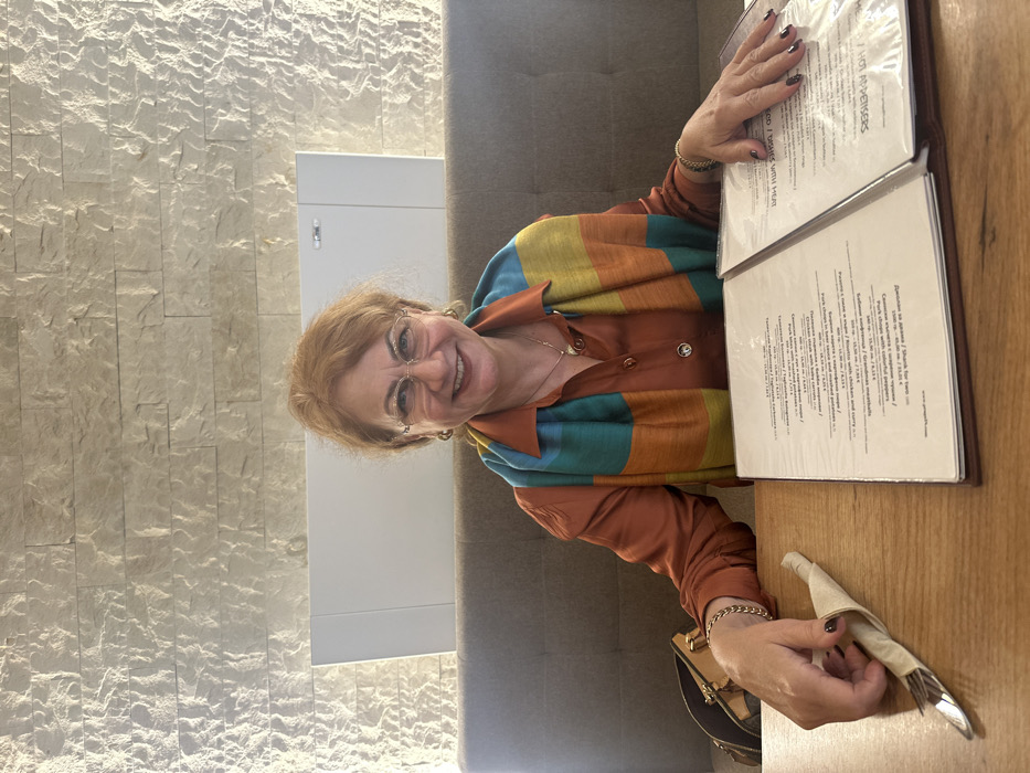 An interior scene depicts a woman seated at a light brown wooden table within what appears to be a restaurant or cafe in Sofia, Bulgaria. The woman, middle-aged with reddish-brown hair, fair complexion, and gold-rimmed oval eyeglasses, smiles directly at the camera. She wears a rust-colored long-sleeved shirt, a horizontally striped scarf (teal, orange, yellow, blue), small gold hoop earrings, a gold chain necklace with a small pendant, a gold-colored chain bracelet on her left wrist, and a dark-colored bracelet on her right wrist. Her fingernails are painted a dark plum or red shade. Her left hand rests on the table near a folded beige napkin containing silverware. Her right hand rests on the right page of an open bifold menu, which displays text in Bulgarian (e.g., "ТЕЛЕШКО," "СВИНСКО") and English, along with prices in Bulgarian Lev (лв). A partially visible brown patterned handbag is positioned on the table to her left. Behind the woman, a light gray upholstered bench extends along the wall. The wall above the bench features a textured, light-colored stacked stone or tile pattern, below which a white rectangular panel or vent is mounted.