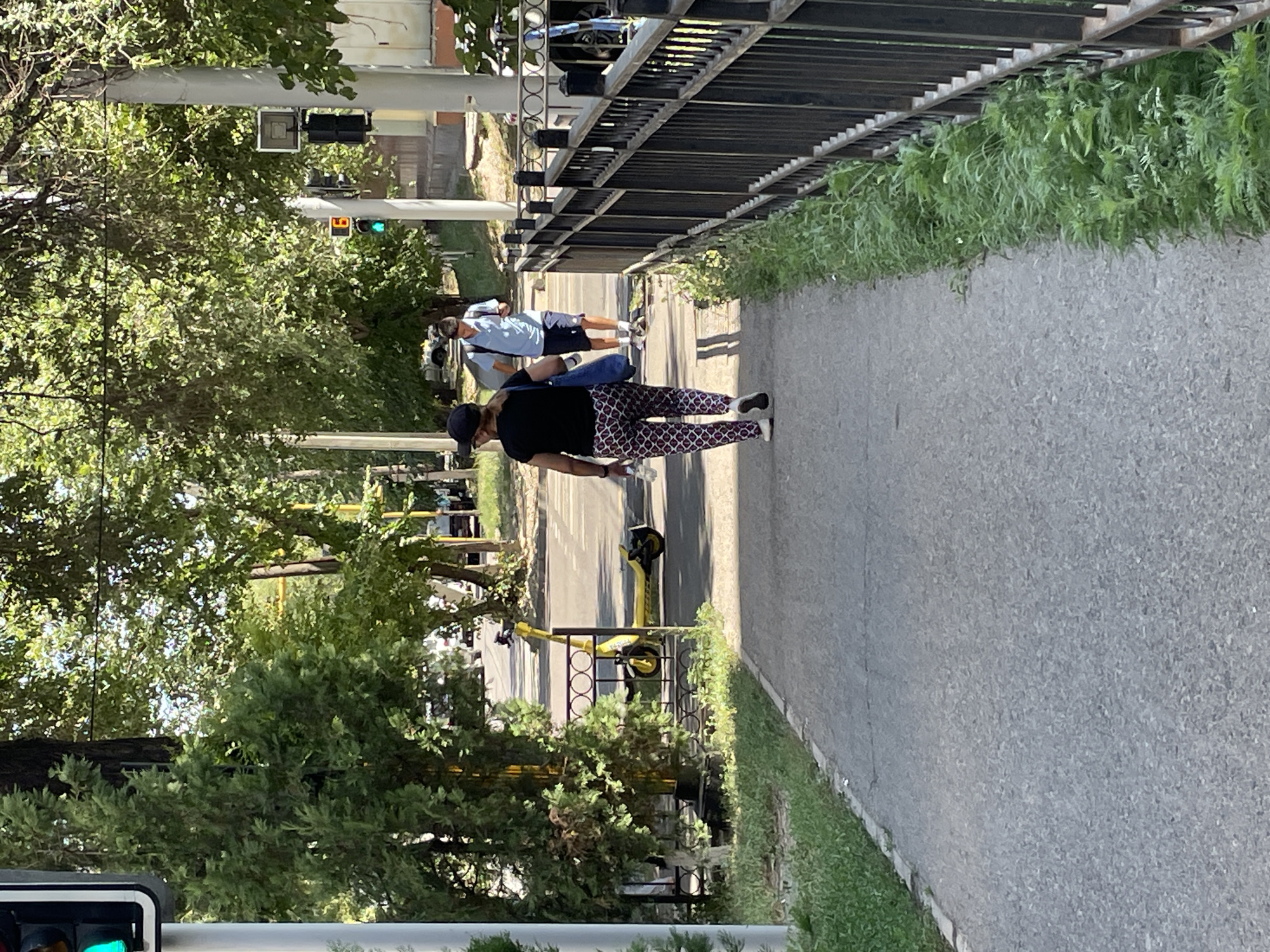 The image captures a street scene in Almaty, Kazakhstan. In the foreground, a paved pedestrian walkway runs parallel to a black metal fence with circular decorative elements. On the walkway, a woman wearing a black t-shirt, patterned leggings, and a black baseball cap is walking away from the camera, carrying a blue bag and a bottle of water. Further ahead on the walkway, a young man in a light blue polo shirt and dark shorts is walking towards the viewer. A yellow electric scooter is parked to the left of the walkway, behind a small black metal fence. Beyond the walkway and fence, trees with dense green foliage line the street. A traffic light with a green signal is visible on the left side of the image, and another traffic light with a green signal and a digital display showing "6" is visible further down the street on the right. The scene appears to be a sunny day in an urban environment.